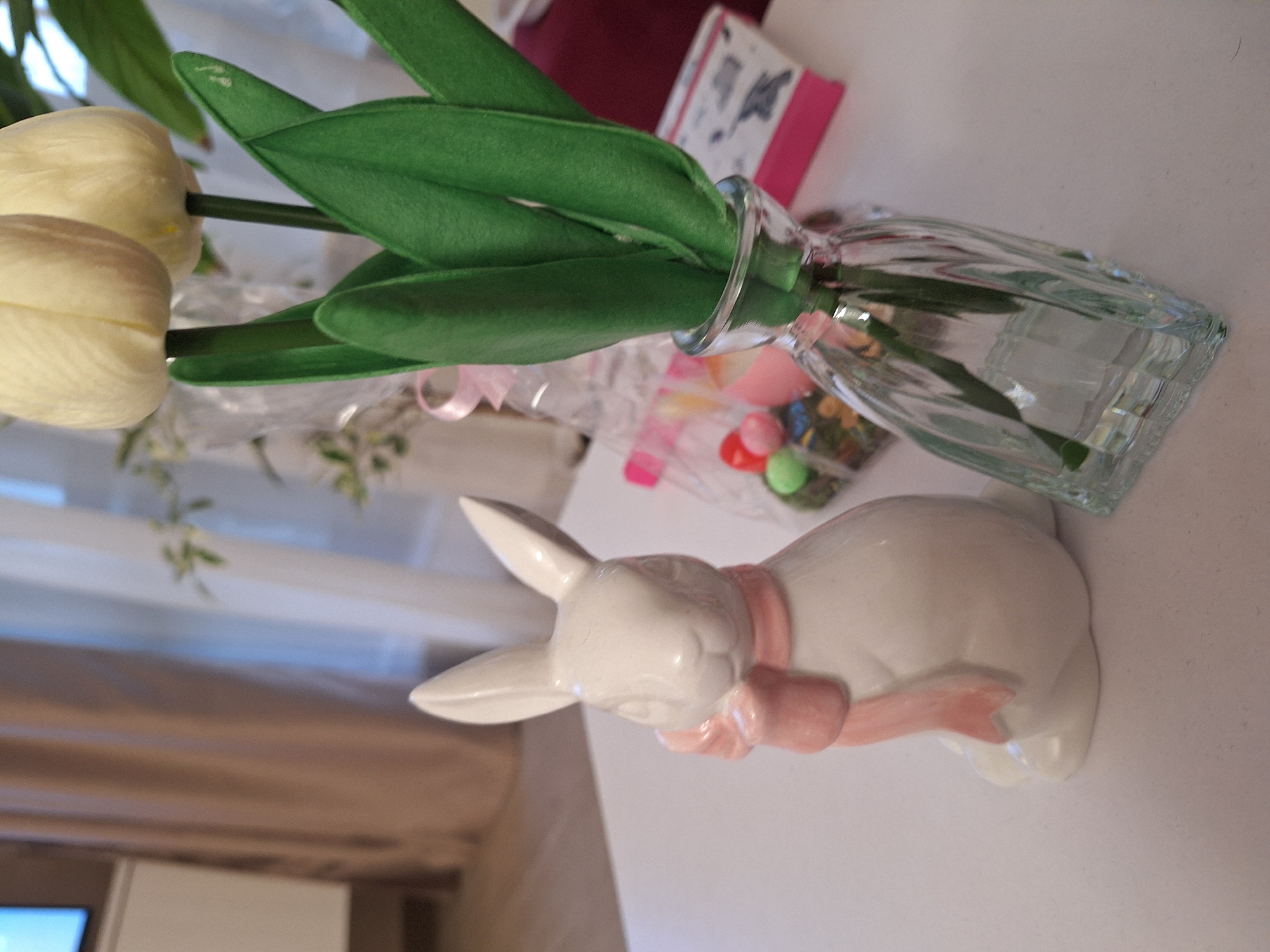 A slightly high-angle, close-up shot shows a white ceramic rabbit with a pink ribbon around its neck, placed on a white surface next to a rectangular clear glass vase holding artificial tulips with green leaves. The stems of the tulips are green, and the flower buds are white. A pink box with a drawing on it and a clear plastic bag filled with pastel-colored candies and plastic grass are in the background. A blurred curtain and furniture are visible in the background. The location is unknown but could be in Sofia, Bulgaria.