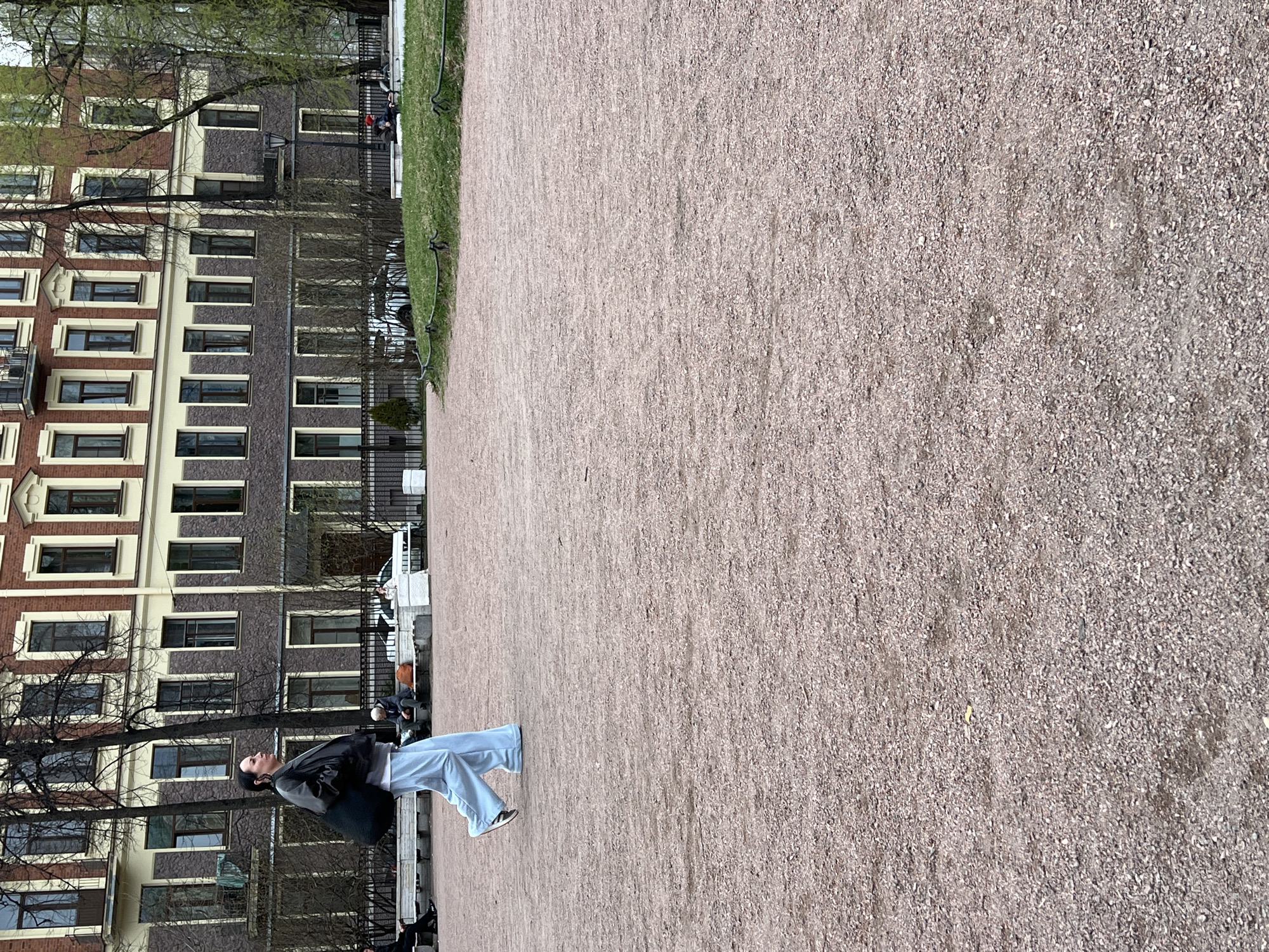 The image shows a gravel path adjacent to a building and a grassy area, likely in Saint Petersburg, Russia. A woman is walking on the path. The building is multi-storied with a combination of red brick and gray facades. A black metal fence separates the path from the grass. Other people are visible near the building.