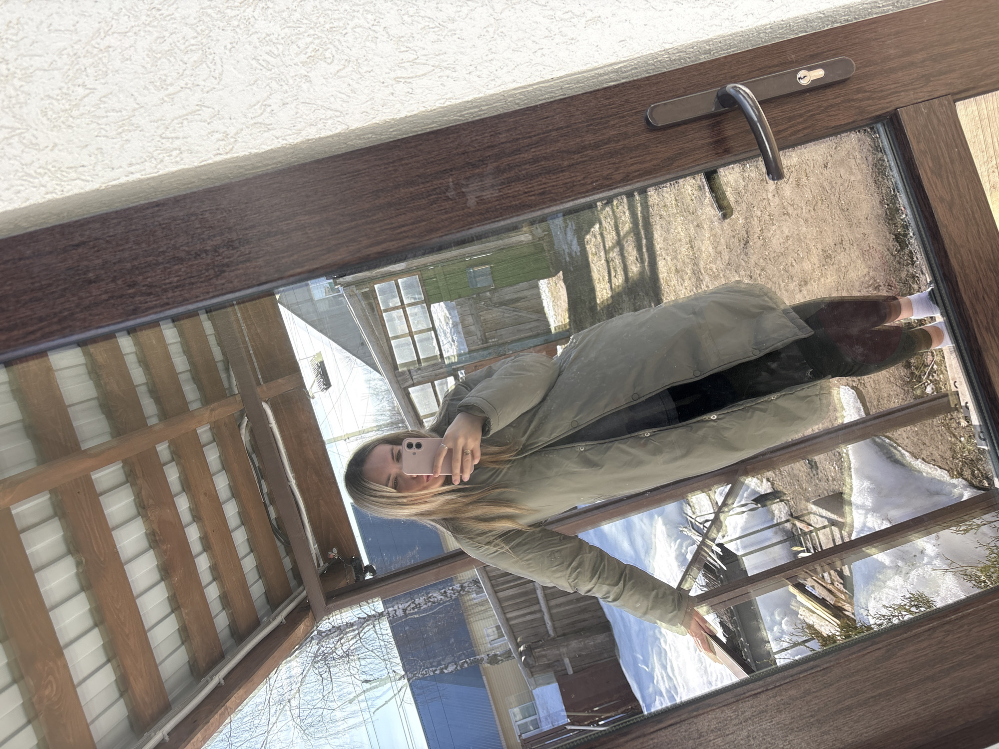 A blonde female, estimated to be in her mid-20s to early 30s, takes a mirror selfie in Votkinsk, Russia. She stands indoors, her reflection captured in a large glass door or window featuring a dark brown frame and a dark handle with a keyhole on the right side. She is dressed in a long, olive-green puffer coat over dark leggings and white socks with dark footwear. Her left hand rests on the brown frame while she holds a pink smartphone to her face with her right hand, positioned to capture the reflection. She wears rings on her right hand.
The outdoor environment, reflected in the glass, indicates a late winter or early spring setting with patches of snow interspersed with bare earth and dry vegetation. A clear blue sky with minimal cloud cover is visible above. The background includes a residential backyard featuring a wooden fence, several leafless deciduous trees (likely birch), and various structures. These structures consist of a house with a light blue corrugated metal roof, another building with a darker roof further back, and a small greenhouse or shed with a dark green door on the right. A wooden overhead canopy or roof structure with wooden beams and a white corrugated metal roof is reflected in the upper portion of the glass. A textured, light-colored wall is visible to the right of the door frame.