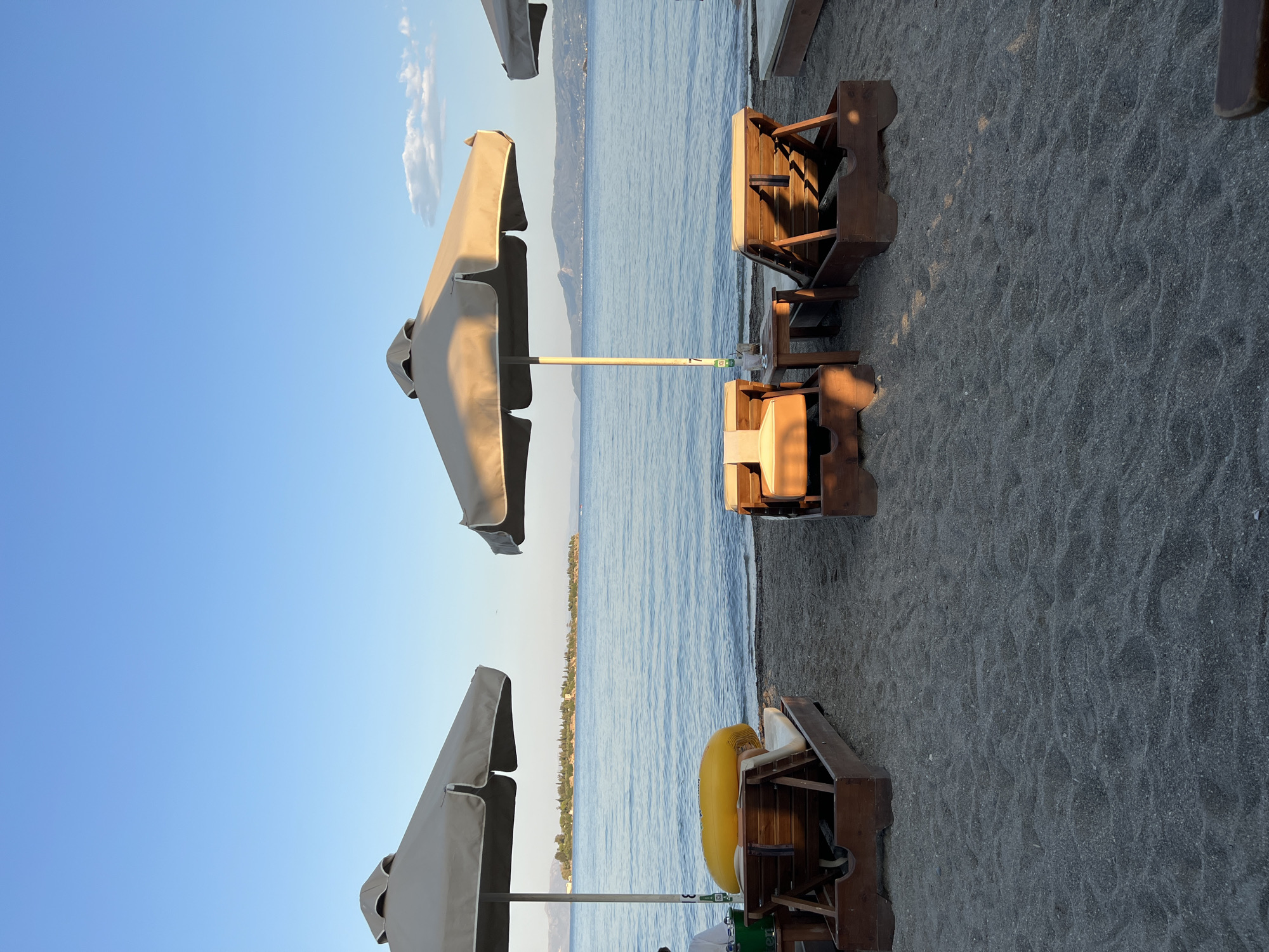 The image depicts a serene beach scene in the city of Eretria, Greece, during the late afternoon or early evening. The foreground is dominated by dark, fine-grained sand with gentle ripples, indicating a recent tide or wind. Several empty wooden sun loungers with beige cushions are arranged on the sand, some partially shaded by large, beige beach umbrellas. To the left, one sun lounger has a bright yellow inflatable raft placed on it. A small wooden table with a drink on it is positioned between two sun loungers in the middle of the frame.
In the midground, the calm, blue sea stretches out to the horizon. The water has a gentle ripple, reflecting the clear, pale blue sky above. A distant, verdant island with scattered trees and buildings is visible across the water, adding a touch of civilization to the natural landscape. The horizon line is marked by the meeting of the sea and the sky, with faint outlines of hills or mountains visible in the far distance.
The lighting suggests a setting sun, casting warm, soft shadows across the sand and the loungers. The overall atmosphere is tranquil and inviting, conveying a sense of relaxation and peace. There are no prominent human figures visible, only a partial view of a person on the far left, sitting on a lounger and partially obscured by an umbrella. The image captures the quiet beauty of a Greek beach at a peaceful time of day.