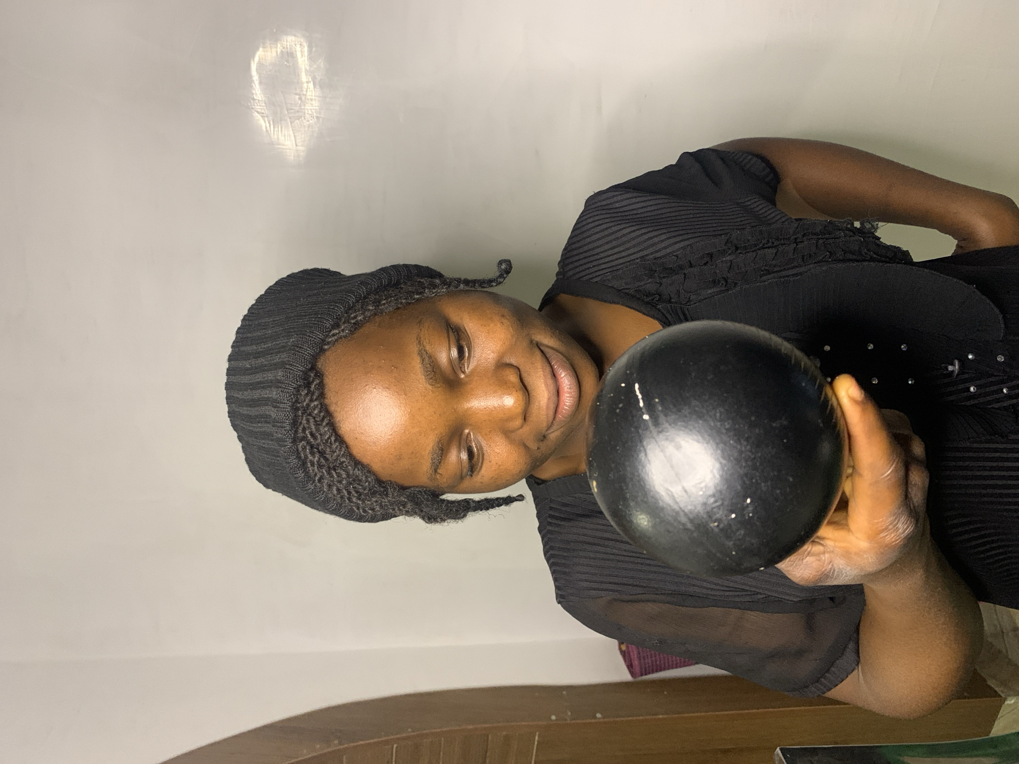 A medium shot captures a young woman of African descent, her face illuminated from above and to the right, as she gazes downwards at a black, spherical object held in her left hand. She is wearing a black knitted beanie that covers her hair, with two braided strands extending from the sides of her head. Her attire consists of a black sheer blouse with ruffled details across the chest, layered over a black garment underneath. The background is a plain, off-white wall with a faint impression of light reflecting from the right. To the left of the frame, a portion of a wooden door with an arched opening is visible. The scene appears to be an indoor setting, possibly a room or studio. The overall impression is one of quiet contemplation, as the woman's attention is fully absorbed by the object she holds. The lighting casts soft shadows, highlighting the contours of her face and the texture of the objects. The image does not contain any specific landmarks or elements that would definitively place it in the city of Jos, Nigeria.