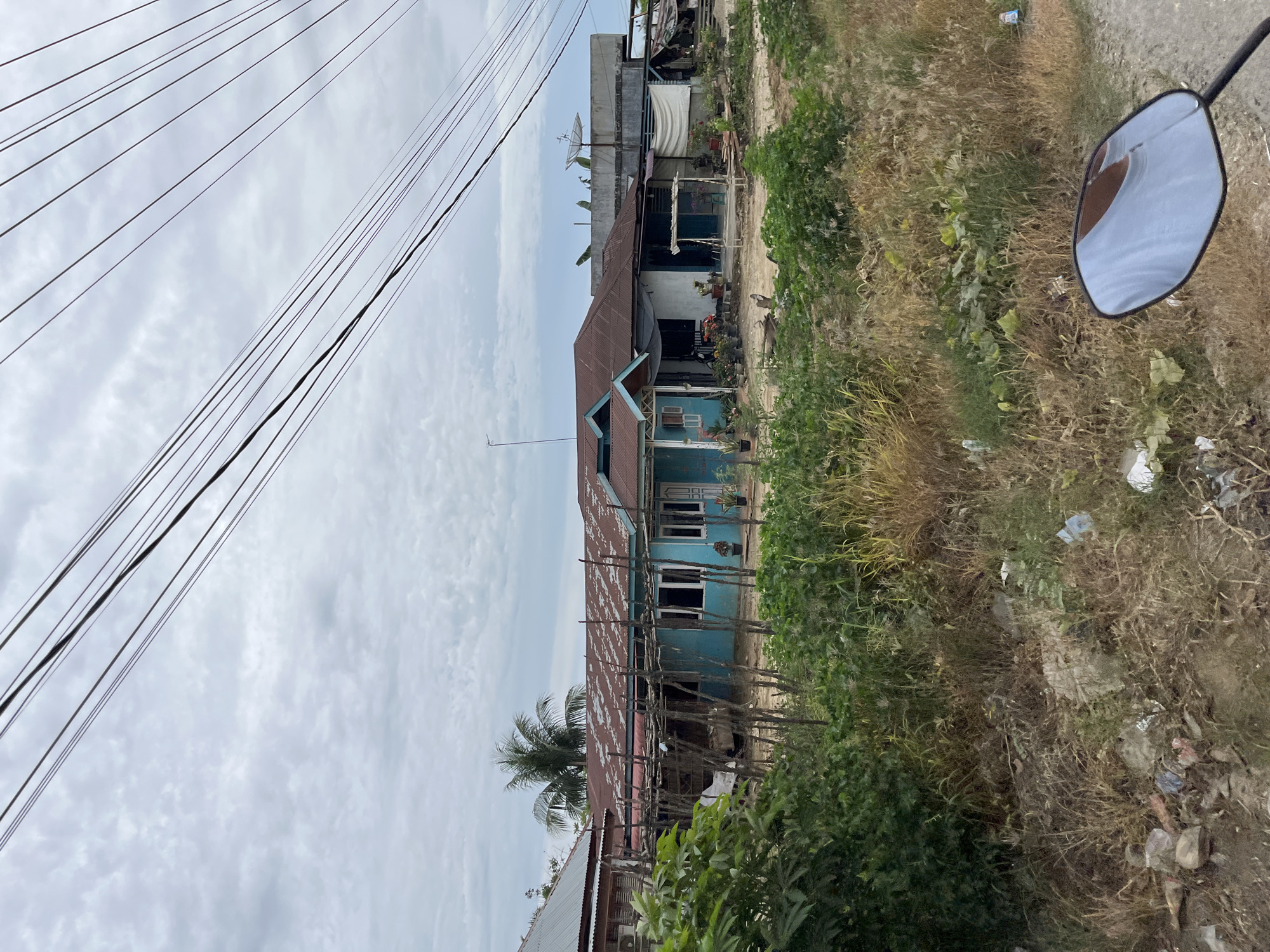 The image depicts a roadside view in Simpangempat, Indonesia. In the mid-ground, several modest buildings with corrugated metal roofs are visible. The most prominent is a light blue house with white trim and dark-paned windows, fronted by a rustic wooden structure. Adjacent to it is another building with a similar tin roof and a white facade. In the foreground, dense, dry vegetation and scattered debris line the edge of a dirt path. Overhead, a network of electrical wires stretches across the cloudy sky. The scene suggests a rural or semi-urban setting, characterized by simple structures and a natural, somewhat unkempt environment.