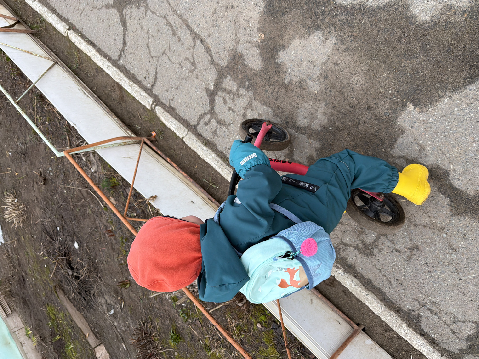 A child in Chaikovsky, Russia, sits astride a red balance bike on a cracked asphalt surface adjacent to a raised, painted concrete curb. The child wears a teal jacket, teal pants, a bright orange hat, yellow rubber boots, and a light blue backpack with a pink pompom. The bike's front wheel is on the asphalt, while the rear wheel is on the concrete. The child is close to a rusty metal railing that surrounds a patch of dirt with some sparse vegetation and remnants of snow.