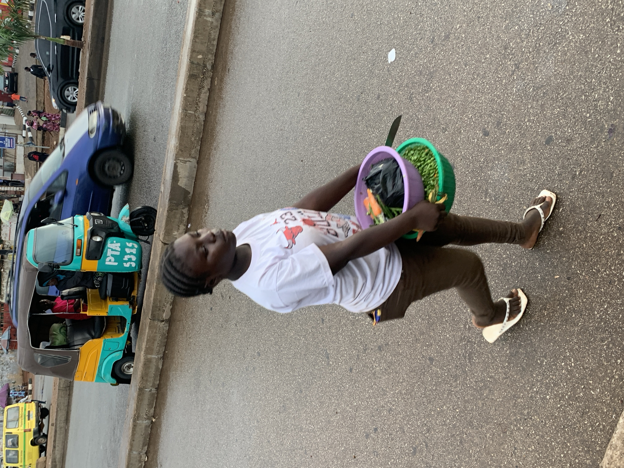 A young Black woman walks on a wet asphalt street in Jos, Nigeria, carrying two bowls of produce. She is wearing a white t-shirt with a graphic of a bull and the number 23, brown leggings, and white flip-flops. The bowls she carries contain carrots, peas, and other vegetables, with a black plastic bag and a knife nestled within. To her left, a turquoise and yellow auto-rickshaw, identified by the license plate "PTA 5315," is parked. In the background, a yellow bus and other vehicles, including a dark SUV and a blue minivan, are visible on the road. The street is lined with a concrete barrier.