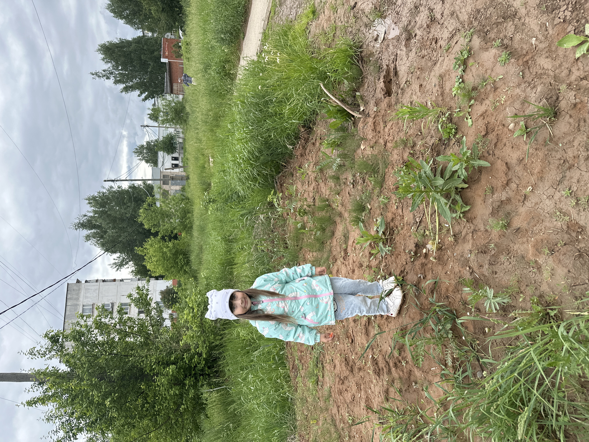 The image depicts a young girl standing in a grassy, partially dirt-covered area in what appears to be a residential neighborhood. The girl is wearing a light-colored unicorn-themed jacket, light blue jeans, and a white hat. She is standing upright and looking toward the camera.
The environment consists of a mix of grassy vegetation and exposed dirt. In the background, several buildings are visible, including a multi-story apartment building. Electrical wires stretch across the sky. The image appears to be taken during the daytime under cloudy conditions.
Given the location context provided, it appears that the scene could be within the city of Chaikovsky, Russia, however, there are no particular identifiable architectural details of Chaikovsky, Russia from the image provided.