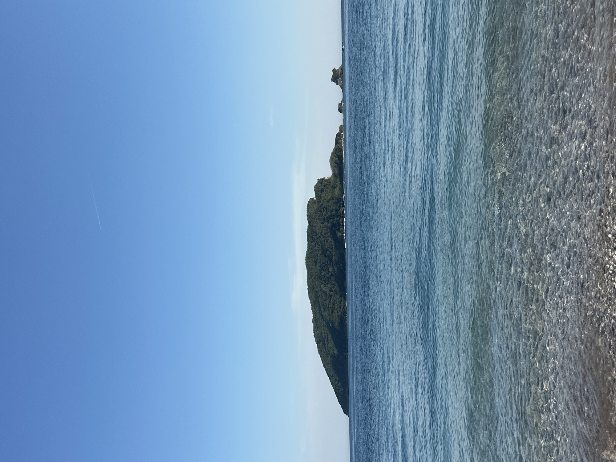 The image depicts a coastal view near Budva, Montenegro. A clear, blue sky occupies the left portion of the frame. An island covered with dense green vegetation is located in the middle of the frame. The right side of the image is occupied by the blue water of the Adriatic Sea. The water is shallow and transparent near the shore. Ripples and subtle wave patterns are visible on the water's surface.