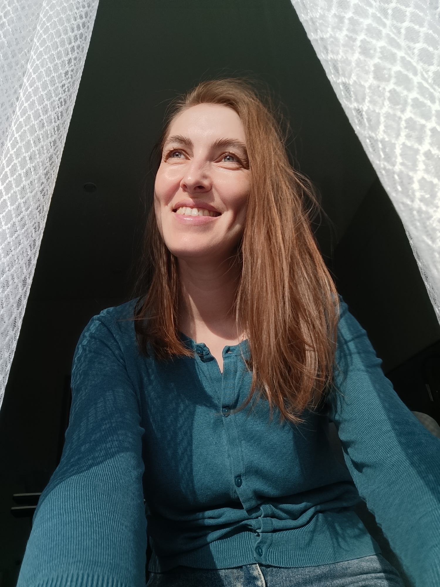 A woman is positioned between white, patterned curtains, looking upwards with a smile. She has fair skin, blue eyes, and auburn hair that falls to her shoulders. She's wearing a teal, buttoned cardigan. The background behind the curtains appears to be dimly lit. The location is Chaikovsky, Russia.