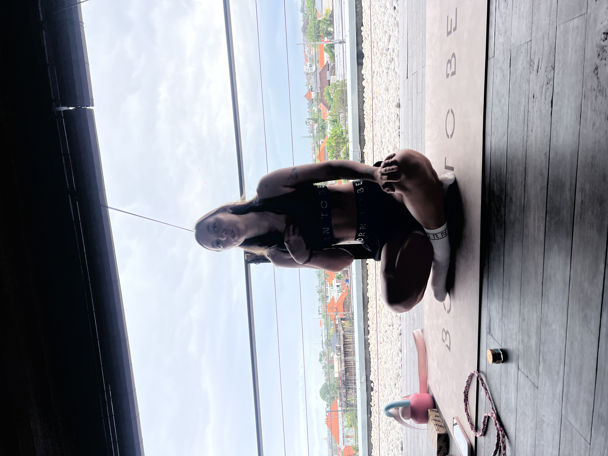A woman is sitting on a yoga mat indoors. She wears a black sports bra and shorts with white socks. The yoga mat is on a wooden floor, with white pebbles lining the edge of the room. Outside the room, the city of Mengwi, Indonesia, is visible, with buildings and trees in the background. There is a window separating the room from the outside. In front of the yoga mat are a yoga block and a rope.