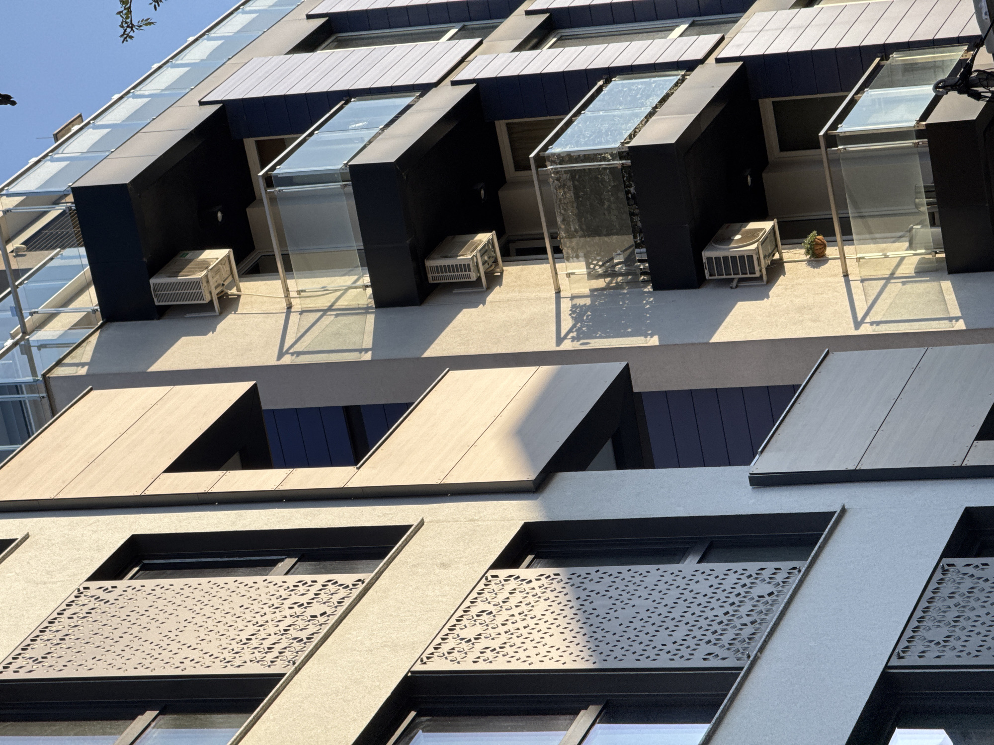 This media file captures a contemporary building facade in Sofia, Bulgaria, viewed from a low angle under bright daylight, indicated by strong shadows. The architecture features a complex interplay of geometric shapes, textures, and materials.
The upper sections of the building display recessed dark panels with angular projections, some of which form open balconies or enclosed glass structures. Several white, rectangular air conditioning units are visible on these ledges, indicating residential or office use. One close-up reveals a small, round, decorative object, possibly a pumpkin or a plant in a pot, next to an AC unit on one of these balconies. The glass elements reflect the clear blue sky and surrounding light.
Below these, the mid-section features flat, light-colored panels that extend outward, some with angular cut-outs, likely serving as sun shades or architectural embellishments. Further down, windows are partially obscured by decorative, laser-cut metal screens with intricate geometric patterns, providing both aesthetic appeal and potentially privacy or sun protection.
The building's color palette is muted, primarily consisting of light gray or beige for the main walls, dark gray or black for the recessed sections and window frames, and the light patterned panels. No visible text is discernible on the building or objects. The overall impression is one of modern, angular design.