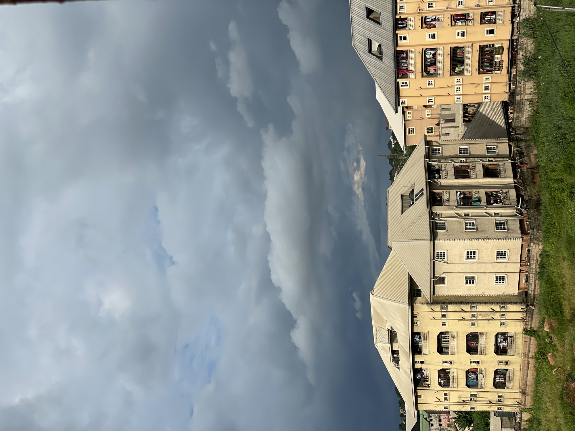 In Nkpor, Nigeria, three apartment buildings dominate the foreground under a dramatic, cloudy sky. The building on the left, painted a light yellow, features multiple balconies adorned with laundry hanging to dry. Its roof is a light grey metal. Adjacent to it, a taller building, also light yellow, displays similar balcony laundry arrangements. This building is connected by a walkway to a third building on the right, which is a lighter shade of beige and has a corrugated metal roof. The buildings are situated on a slightly elevated grassy area, with visible rocks in the foreground. The sky is filled with a mix of dark, heavy clouds and lighter, scattered ones, suggesting potential rain. The scene is devoid of visible people, but the presence of drying laundry indicates recent human activity. The overall impression is one of a residential urban environment under a dynamic, cloudy sky.