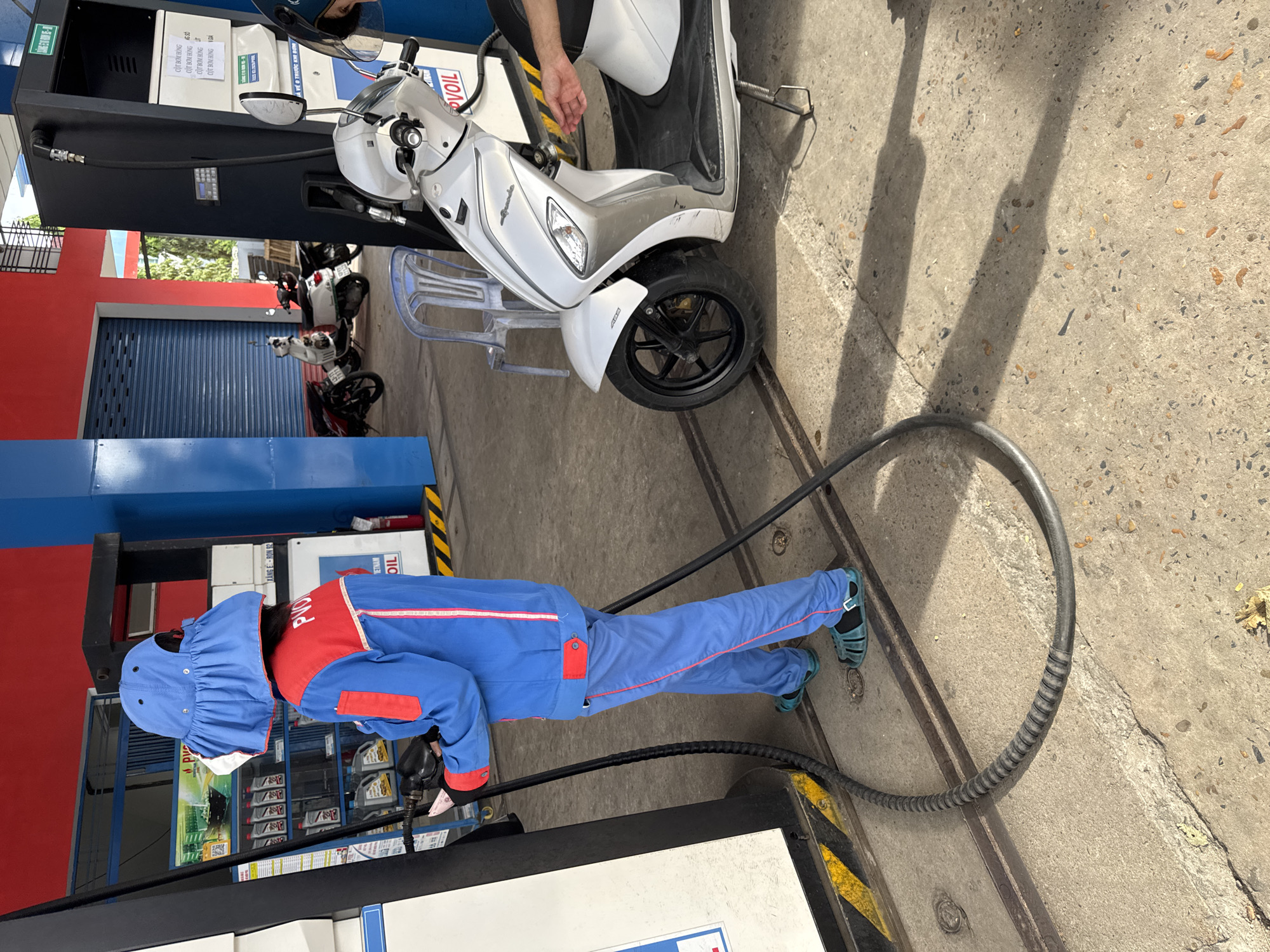 At a gas station in Unknown, Unknown, an attendant wearing a blue and red uniform and a blue sun hat is seen from behind, dispensing fuel into a white scooter. The attendant is holding a black fuel nozzle connected to a gray hose, which is attached to a gas pump. The scooter is parked next to the pump, and its rider's hand is visible near the fuel tank. Several other scooters are parked in the background. The ground is made of concrete, and the scene is illuminated by natural sunlight, casting shadows on the ground. The gas station features red and blue accents on its building.