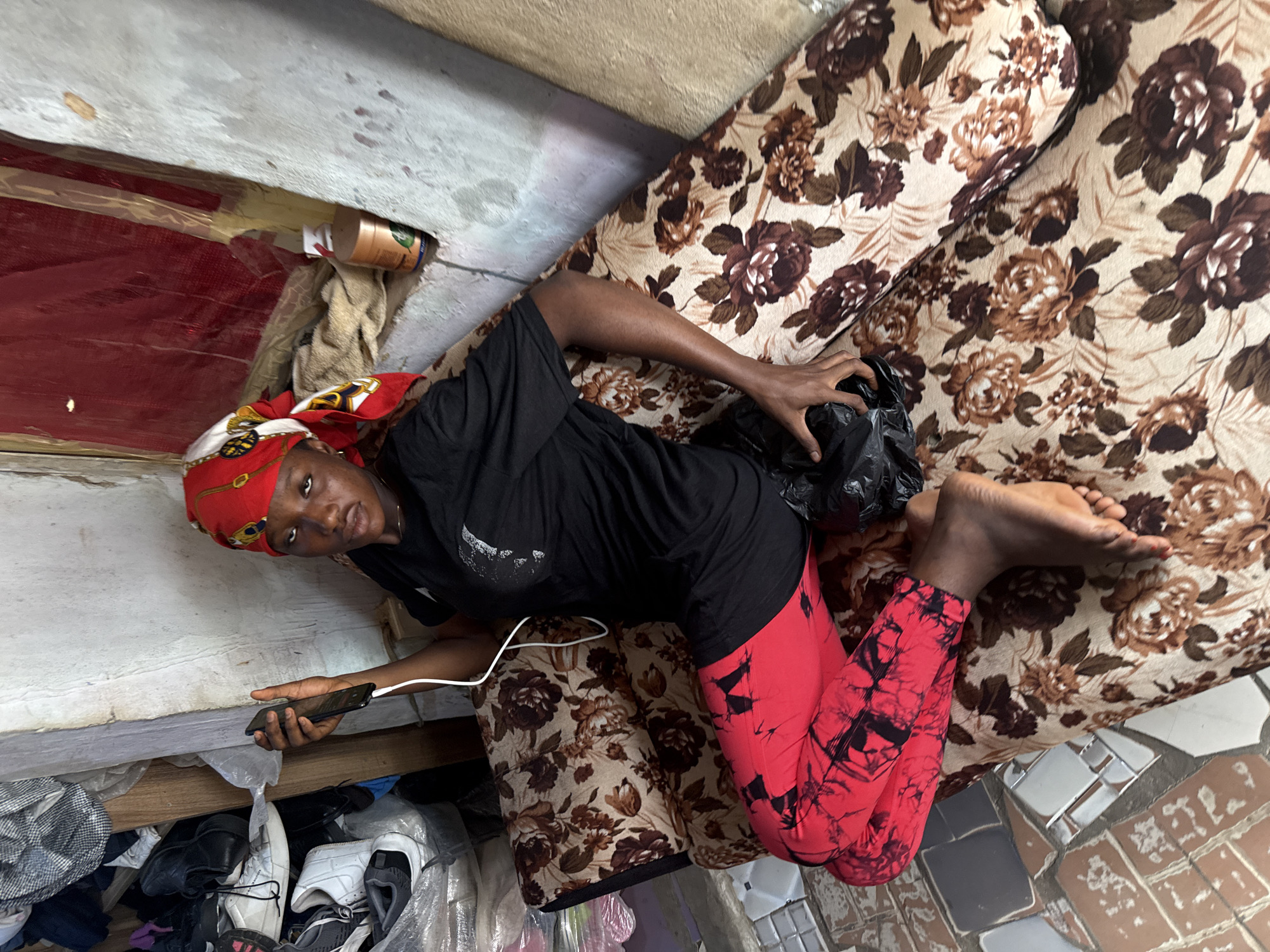 A young woman, appearing to be in her late teens or early twenties, is seated on a floral-patterned couch in what appears to be a dimly lit room. She is wearing a red and white patterned headscarf, a black t-shirt, and red pants with a black tie-dye pattern. Her left arm is extended, holding a smartphone that is connected to a white charging cable. Her right hand is resting on a black plastic bag on her lap. The couch is positioned in a corner of the room, with a shelf full of various items, including shoes, bags, and clothing, visible behind her to the left. To the right of the couch, a wall with a red, possibly plastic, covering is visible, along with a light-colored cylindrical object on a ledge. The floor is tiled with a mosaic of light and dark squares. The overall impression is that of a humble, cluttered living space, possibly in a residential area of Abuja, Nigeria.