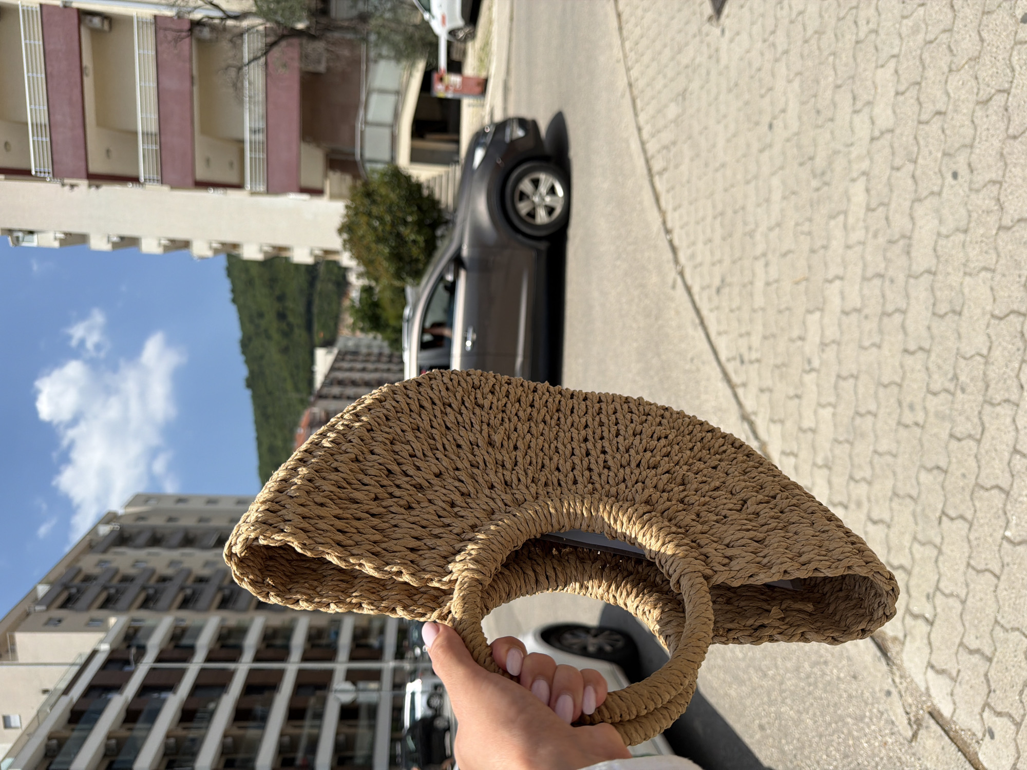 A person's hand with light pink nails holds a woven straw bag in the foreground. The bag is angled and partially obscures a street scene. In the background, there are apartment buildings, a dark-colored car driving on the street, trees, and a green, hilly landscape. The ground in the foreground is paved with interlocking concrete pavers. The sky is blue with some white clouds. The image appears to have been taken on a sunny day in Budva, Montenegro.