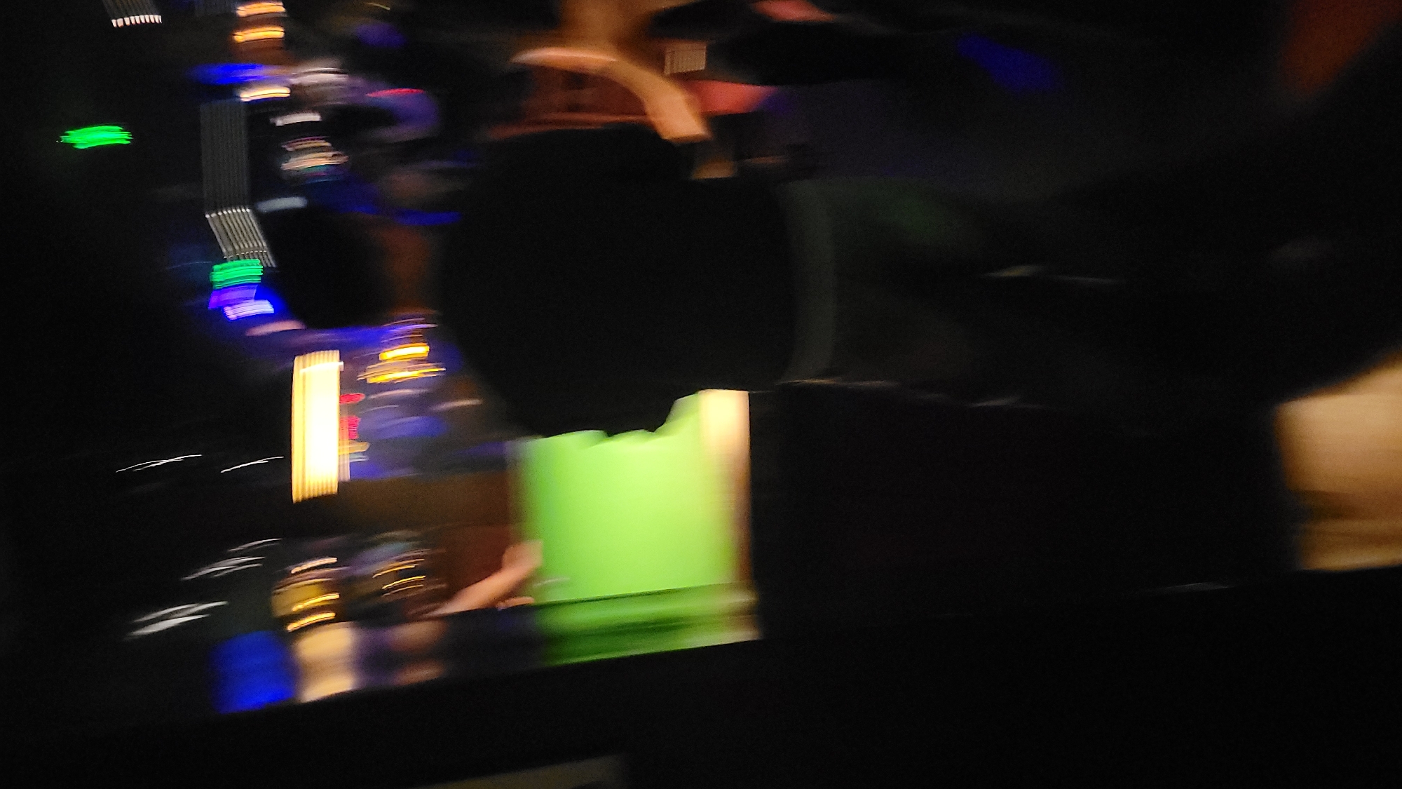 The image depicts a blurry scene at night, possibly taken from a moving vehicle. There are various blurred lights of different colors, including green, blue, yellow, and white, suggesting urban lights or traffic. An out-of-focus dark object is prominent in the foreground, potentially a person or object inside the vehicle. A bright green rectangular shape is also visible, possibly reflecting light from a building or sign. The exact location within the city of Unknown, Unknown is not discernible from the image due to the blurriness and lack of specific landmarks.