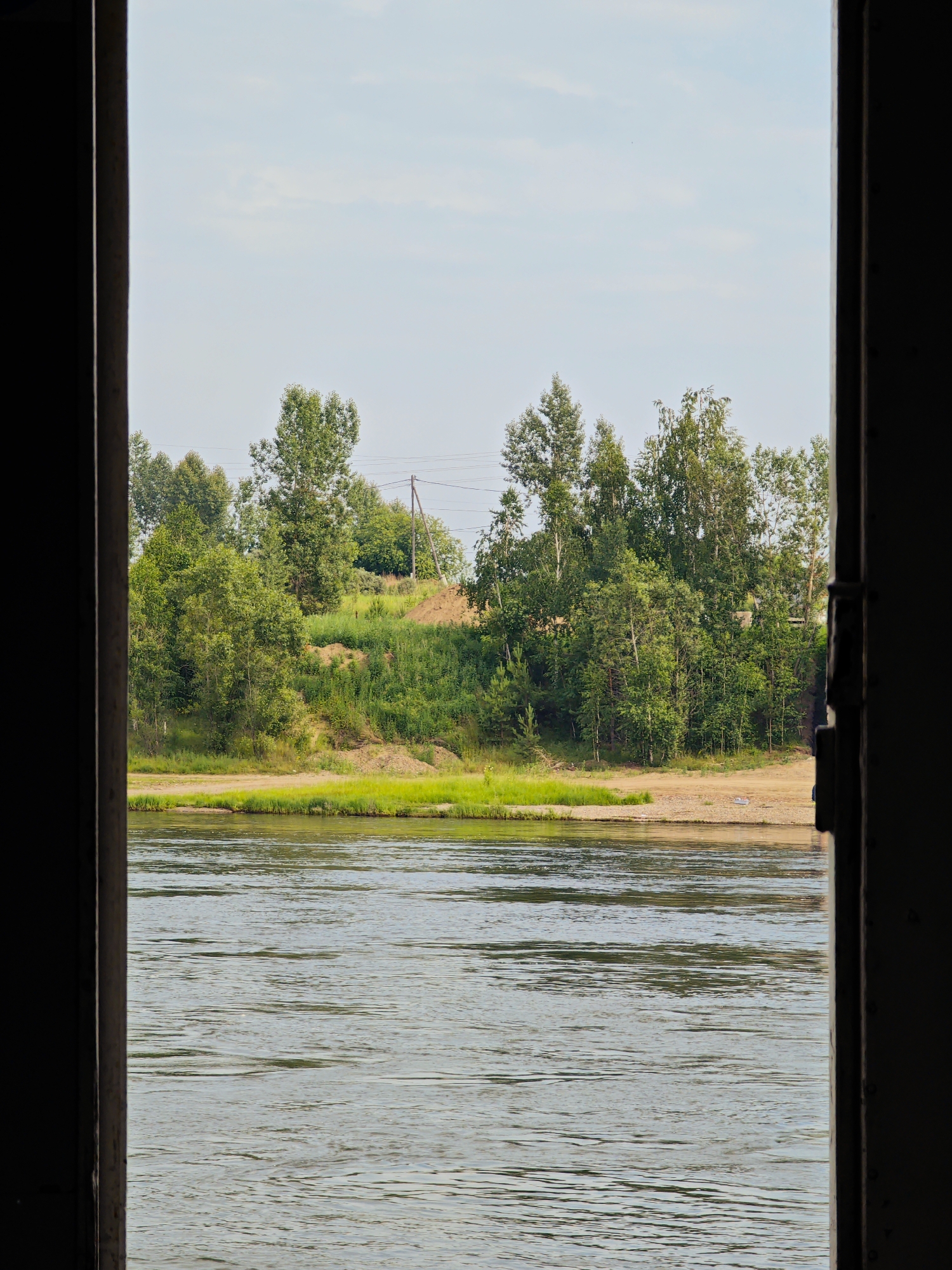 The image shows a view of a wide river from a dark, partially obscured opening, likely a doorway or window frame. The river occupies the lower half of the frame, its surface rippling with light reflections. Beyond the river, a sandy bank leads to a lush green embankment covered in dense trees. In the background, a few structures and utility poles are visible amidst the foliage. The sky above is a pale blue with scattered clouds. The scene appears to be set in a natural environment, possibly on the outskirts of a city. The overall impression is one of a tranquil, natural landscape viewed from a sheltered vantage point. The specific location in Sosnovoborsk, Russia, cannot be determined from the visual information provided in the image alone.
