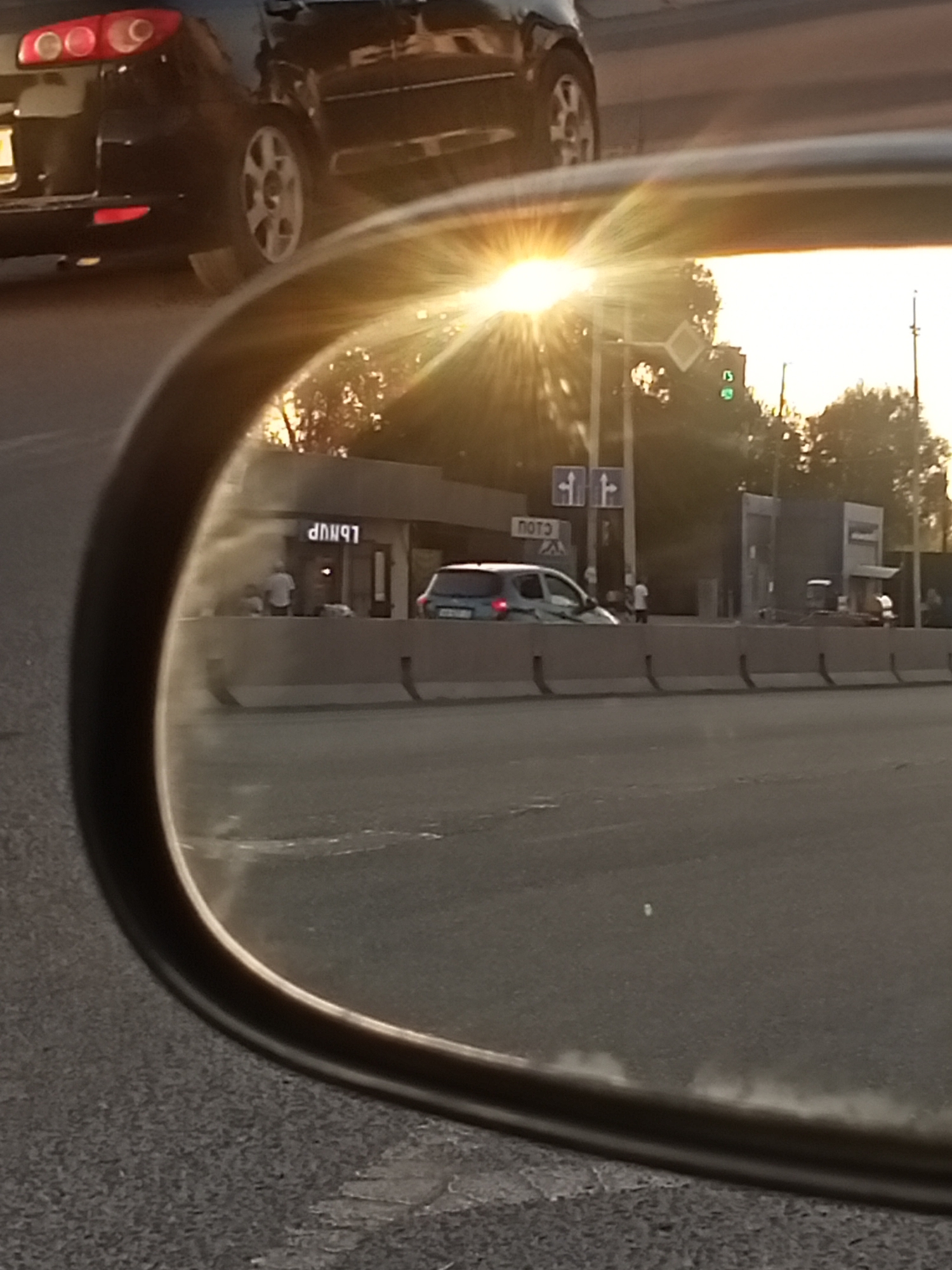 In Sokuluk, Kyrgyzstan, a reflection in a car's side-view mirror shows a street scene at dusk. The sun is low on the horizon, casting a bright glare and lens flare in the sky. In the foreground of the reflection, a silver hatchback is parked in front of a building with illuminated signage. The signage appears to be in Cyrillic script. A person in a white shirt stands near the building's entrance. Behind the silver car, a white van or truck is visible. A concrete barrier separates the road from the sidewalk area where the building is located. In the background of the reflection, traffic lights are visible, with one showing a green signal. Two blue directional signs are also present. Further in the distance, trees and utility poles can be seen. The original image shows a portion of the car the mirror is attached to, with its taillights visible, and the road surface.