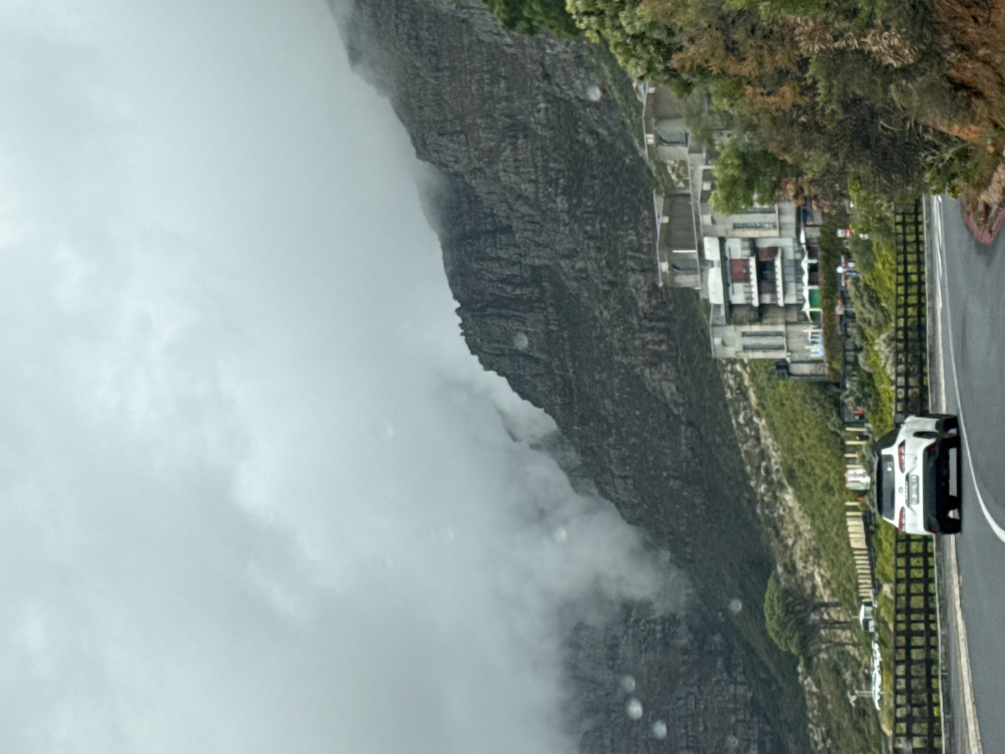 The image depicts a view of the Table Mountain in Cape Town, South Africa, shrouded in clouds. A road runs along the base of the mountain, with several parked cars. Buildings are visible nestled on the hillside, among dense trees and vegetation. The sky is overcast. The angle of the photo indicates the camera was positioned at an unusual angle, approximately rotated 90 degrees.