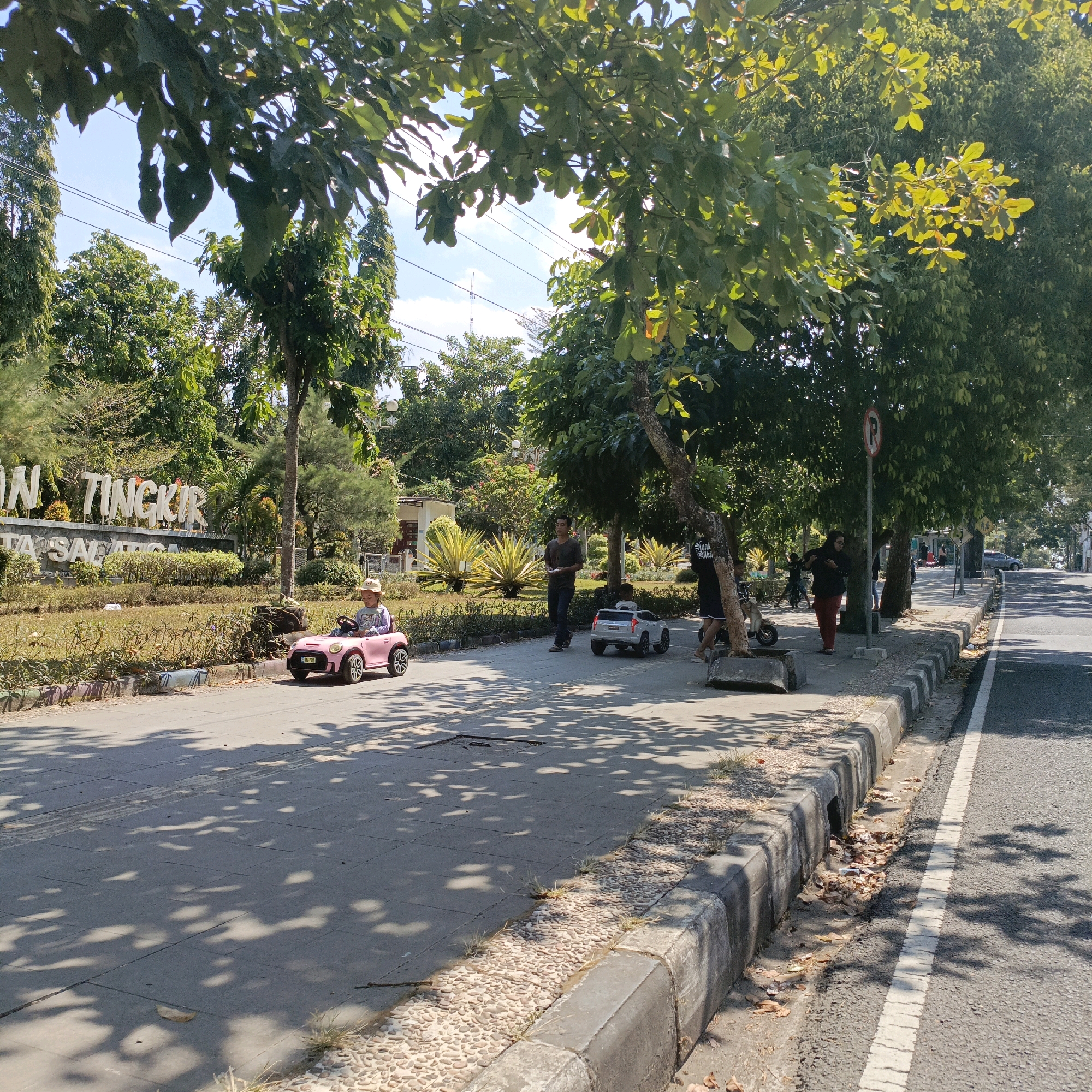 A child is riding a pink toy car on a paved pathway in front of a landscaped area with a sign that reads "Tingkir". The pathway is adjacent to a road. Several trees provide shade on the pathway, casting dappled shadows. Another child is riding a white toy car further down the pathway. A man is walking on the pathway behind the children. In the background, there are more trees and buildings. The weather appears to be sunny.