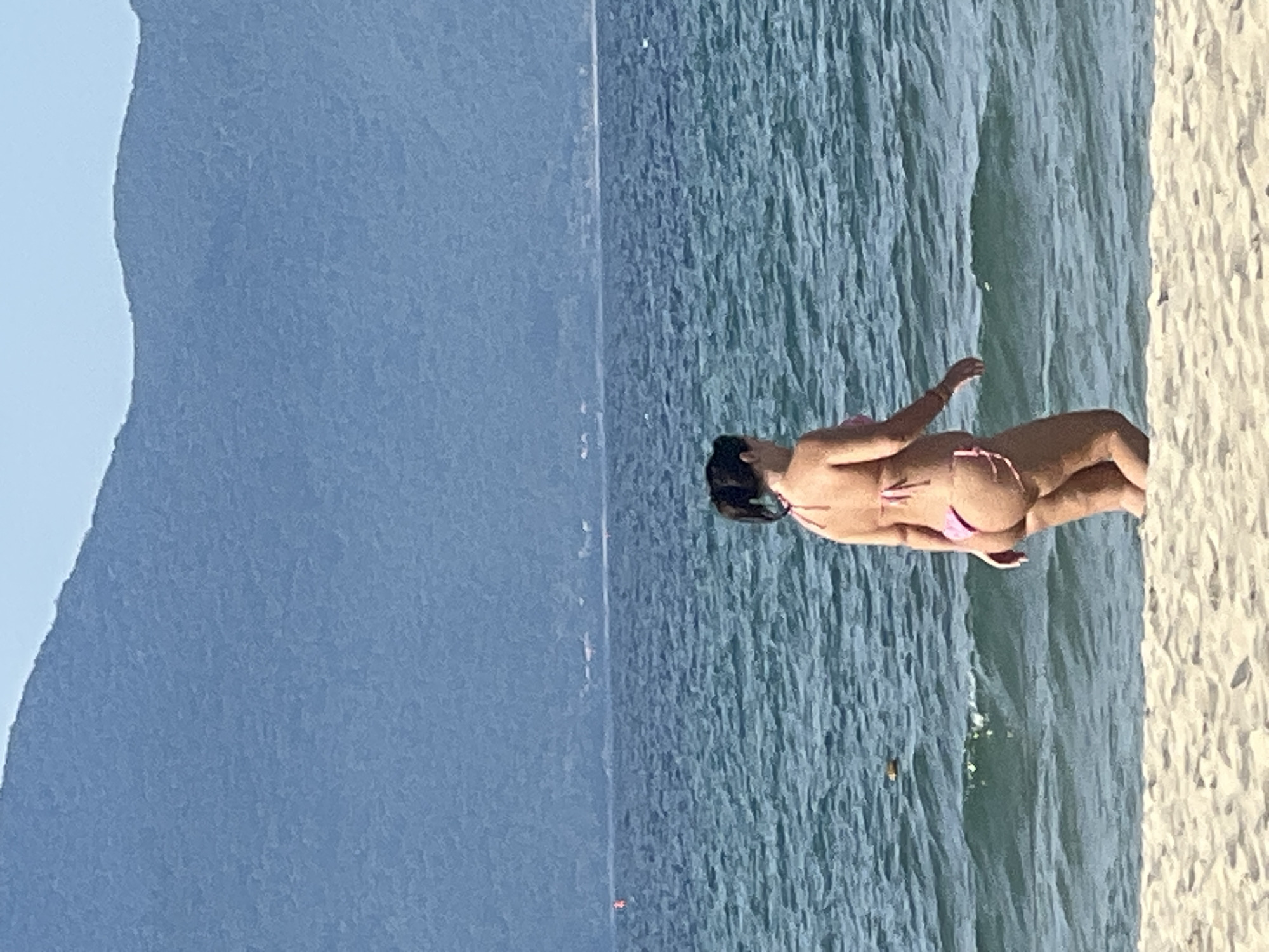 A woman in a pink bikini walks in the shallow water at a beach. She is facing away from the camera, with her back to the viewer. The water is blue and choppy, with small waves breaking on the shore. In the background, there are distant mountains under a clear sky. The sand in the foreground is light beige. The location is My Khe Beach in Da Nang, Vietnam.