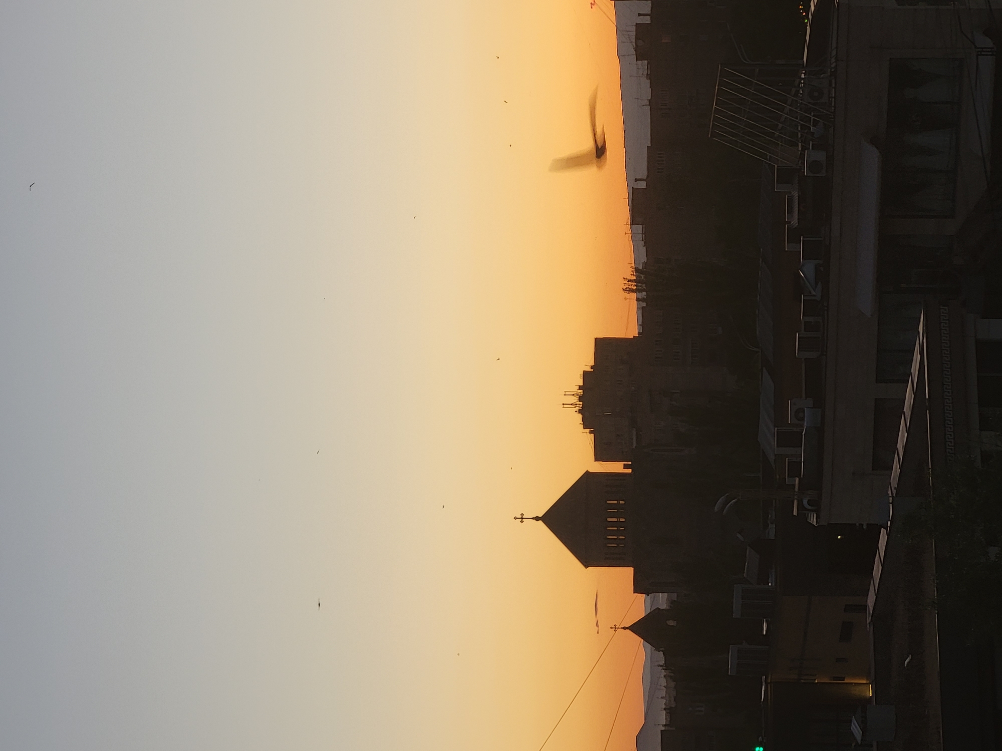 The image depicts a sunset view of Yerevan, Armenia. The sky transitions from light grey to deep orange. Several birds are visible in flight. Silhouetted against the sunset are buildings, including a church with two steeples and crosses atop them. Modern apartment blocks and commercial buildings are also part of the cityscape. Utility lines cross the sky. The overall scene captures the atmosphere of dusk in the city.