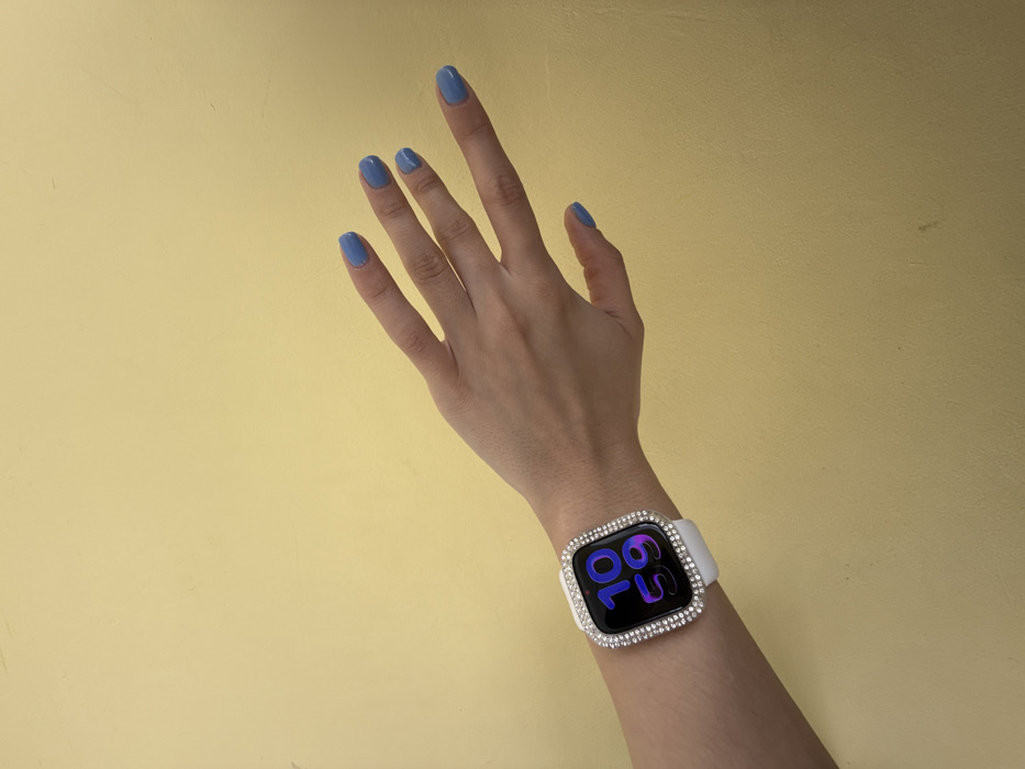A left human hand, with fair skin and fingers slightly splayed, is depicted against a plain, solid yellow-ochre wall in Molodezhnyi, Russia. The fingernails are painted with light blue polish. A smartwatch, identified as an Apple Watch, is worn on the wrist with a white silicone strap. The watch face is enclosed within a clear protective case adorned with two rows of small, clear rhinestones. The watch screen displays the time "10 59" in dark blue digital characters. Minor scuff marks are visible on the yellow wall.