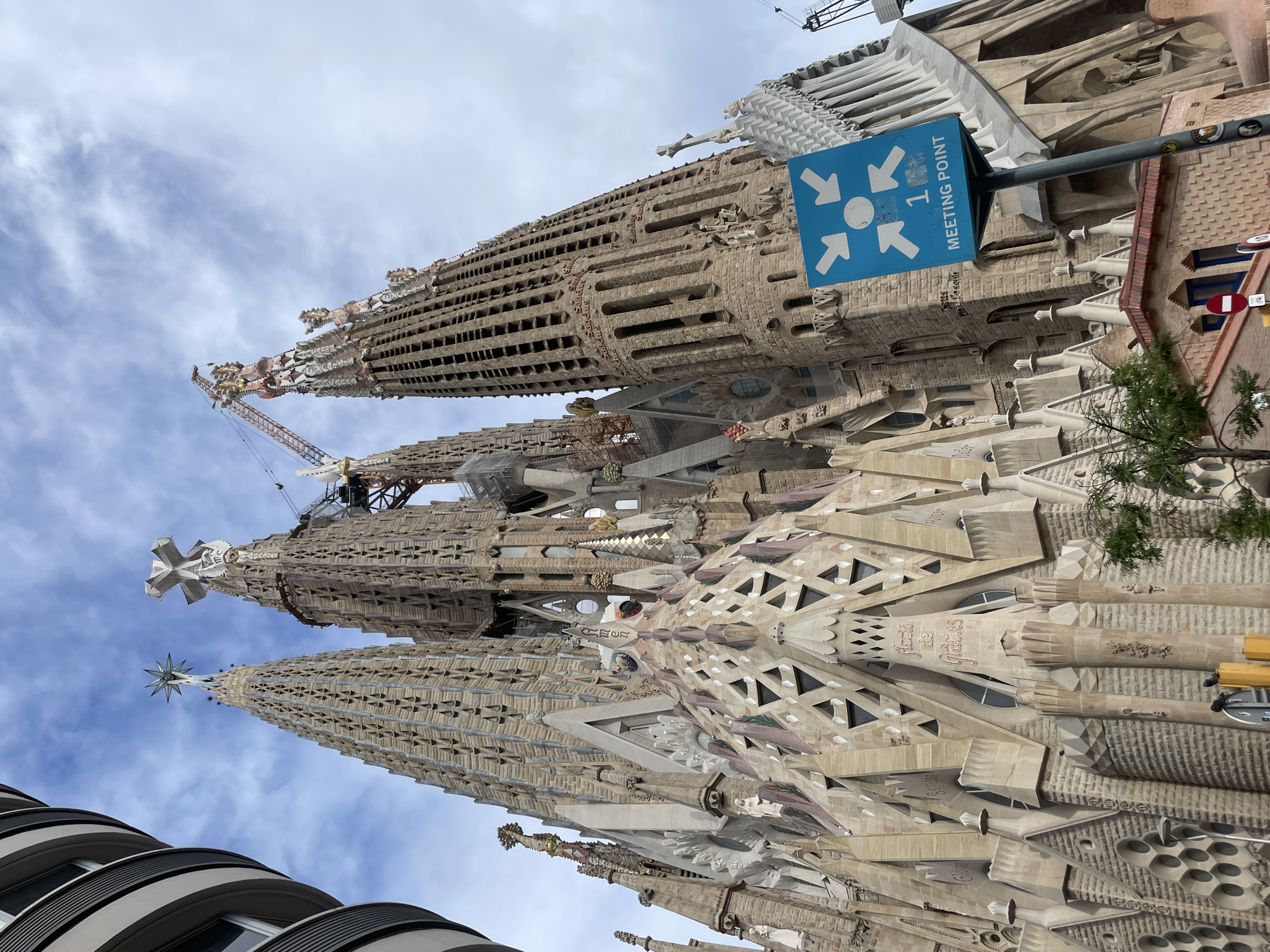 The image shows a low-angle view of the Sagrada Familia basilica in Barcelona, Spain. The basilica's ornate towers dominate the frame, showcasing intricate architectural details and stone carvings. A blue sign with white arrows pointing inward and the words "MEETING POINT 1" is visible, indicating a designated meeting spot. Construction cranes are present, signaling ongoing work on the basilica. The sky is partly cloudy. In the bottom left corner, a portion of a modern building with rounded balconies is visible.