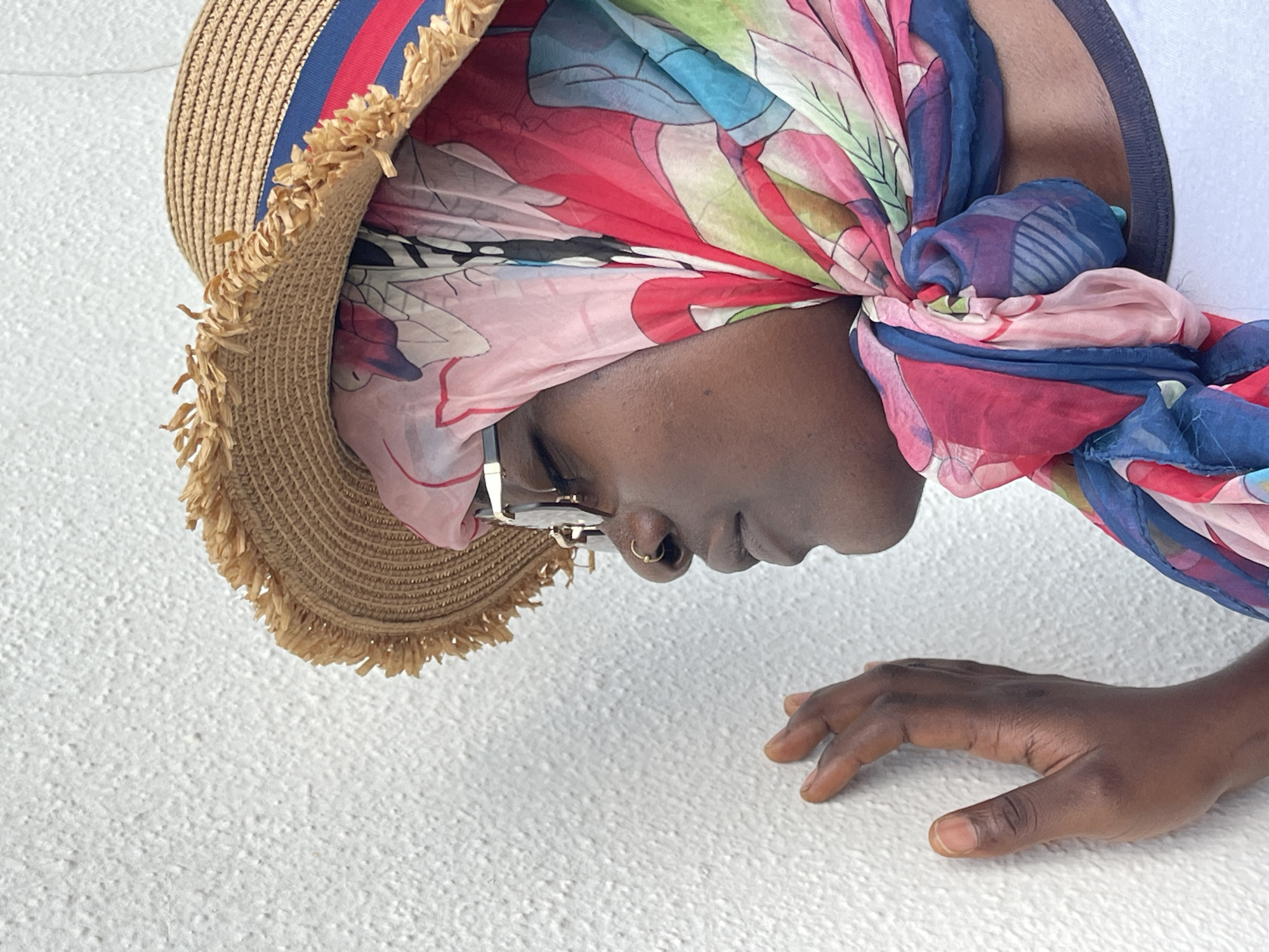 A person wearing a straw hat and colorful headscarf is positioned over a white textured surface. The hat is trimmed with frayed edges and blue and red stripes. The headscarf features a floral print in shades of pink, blue, red, and green. The person wears gold-framed glasses and a nose ring. One hand is visible on the white surface. The location in Gwarinpa, Nigeria, is not discernable from the image.