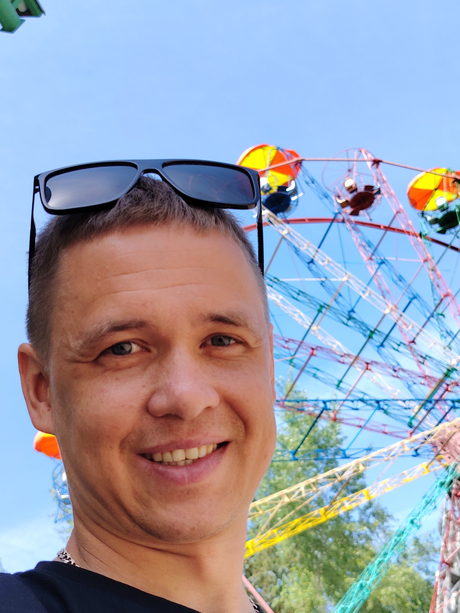 A close-up photograph depicts a smiling man with short hair and sunglasses perched on his head, seemingly taking a selfie. In the background, a vibrant Ferris wheel is visible, suggesting a fairground or amusement park setting. The location is identified as Chaikovsky, Russia, based on associated information. The sky is clear and blue, indicating a sunny day. The man is wearing a dark shirt and a silver chain is visible around his neck. The Ferris wheel has colorful carriages, adding to the festive atmosphere.