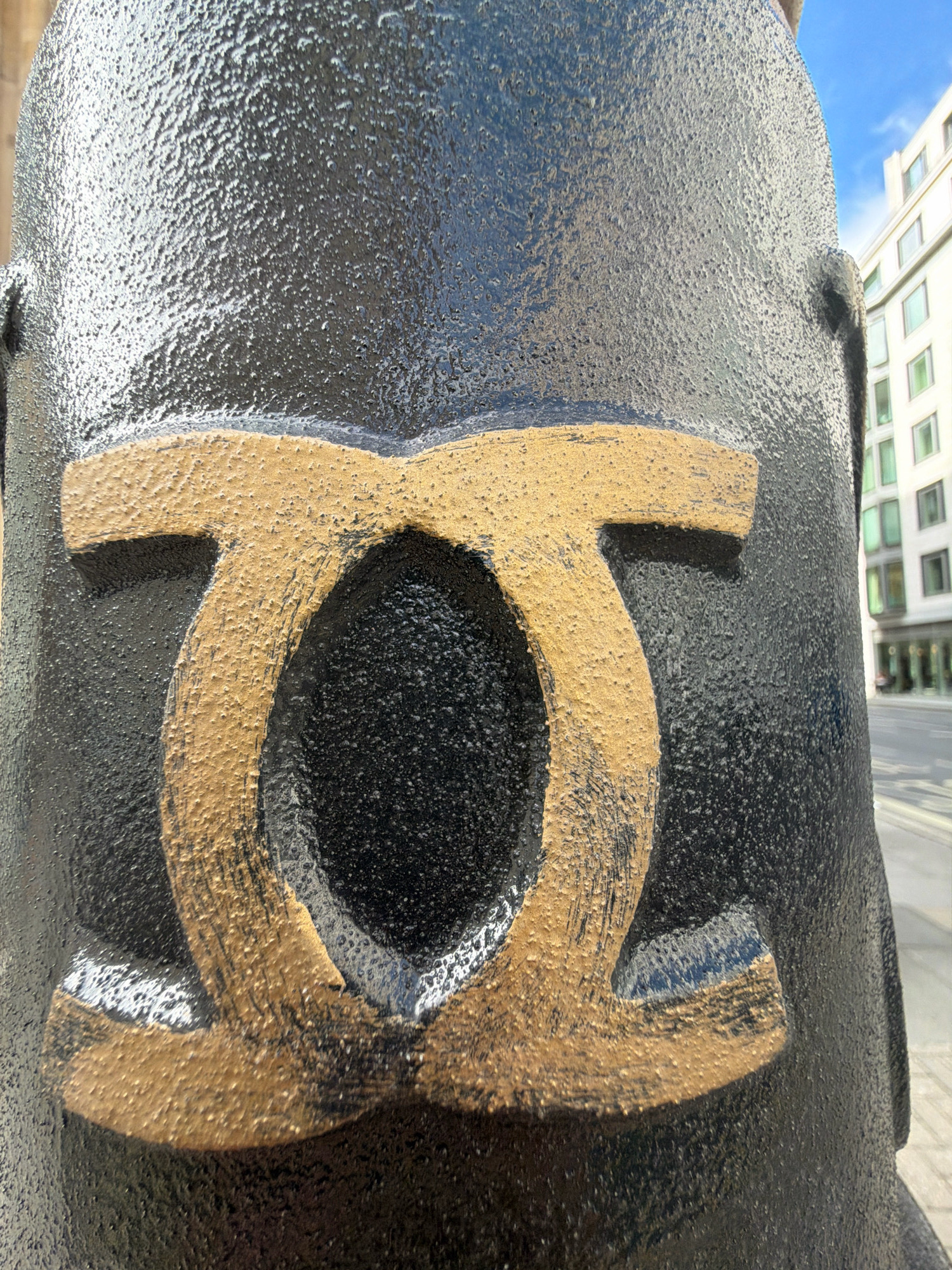 A close-up, eye-level, outdoor shot focuses on a black, textured lamppost with a raised, golden symbol. The symbol is composed of two interlocking oval shapes, resembling an infinity symbol or the Greek letter Omega. The texture of the lamppost is rough and pebbled, with the golden symbol having a slightly rougher, applied texture.
In the blurred background, a modern building with rows of windows is visible, suggesting an urban environment. A street is also faintly visible, with hints of pavement and possibly traffic lights or street signs. The sky above is bright blue with some white clouds.
This lamppost is located on Regent Street in London, United Kingdom, specifically in front of the Cartier store.