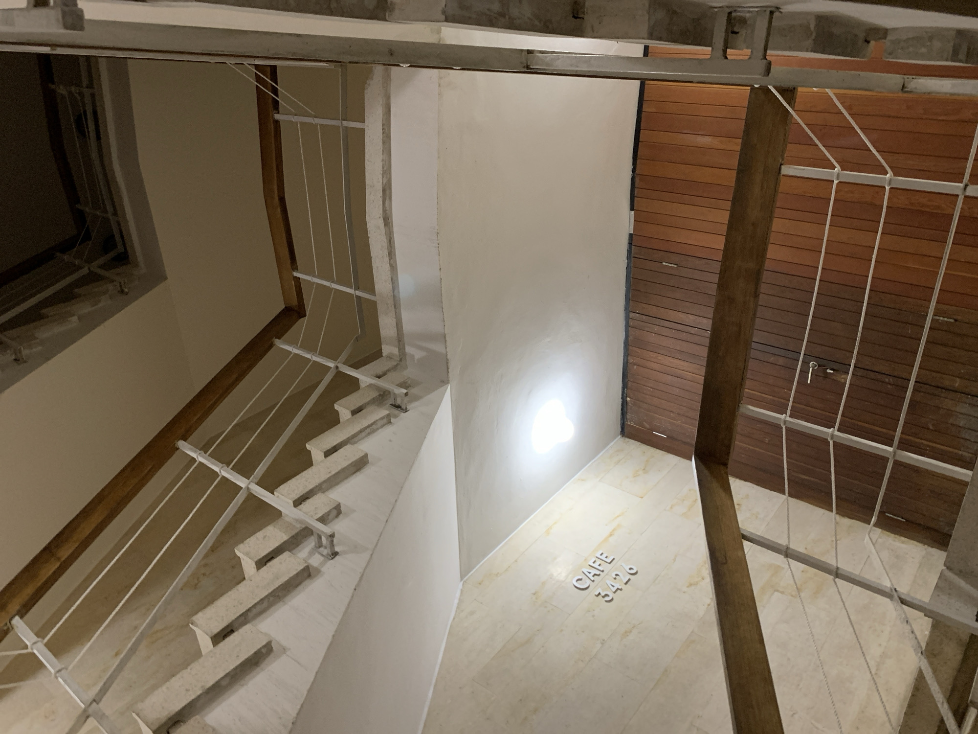 The image displays an interior view of a multi-level staircase within a building in Tel-Aviv, Israel. The vantage point is from an upper floor looking downward across several flights and landings. The stairs are constructed of light-colored, possibly concrete or stone, material. A white metal railing with multiple horizontal rods or cables runs along the staircase, topped with a light brown wooden handrail.
Walls exhibit two distinct finishes: one section is clad in horizontally-oriented, light beige marble or stone tiles, on which white dimensional letters spell "CAFE" above "3426". Another wall section and the ceiling are painted a uniform off-white or light beige color. A single, oval-shaped, white ceiling light fixture provides illumination to a lower landing. At the base of the visible scene, a dark brown wooden paneled door with a silver-toned handle is present, flanked by dark brown wooden paneling. No individuals are visible, and no actions or interactions are depicted.