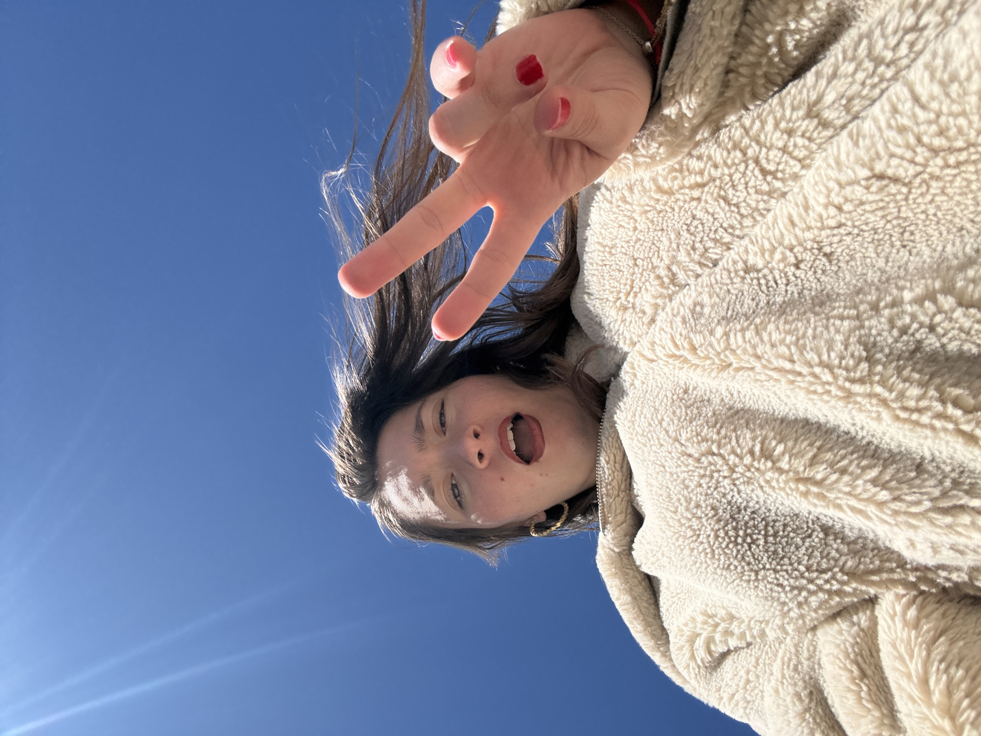 A low-angle shot reveals a young woman against a clear blue sky. She is wearing a cream-colored, fleece-like jacket. Her brown hair is tousled, and she is sticking her tongue out. Her hand is raised, making a peace sign. Her fingernails are painted red. The exact location in Sofia, Bulgaria cannot be determined with the current information.