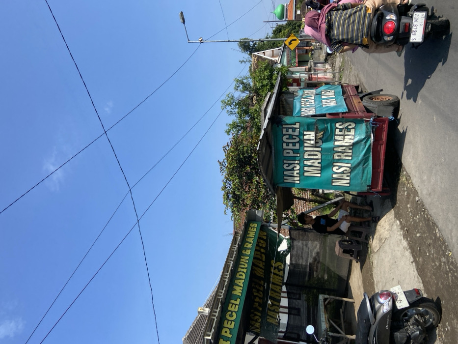 The scene is a street in Mojosari, Indonesia, at latitude -7.504 and longitude 112.552. There is a food cart with a sign that reads "Nasi Pecel Madikun Nasi Rames" in Indonesian. The cart is red with a green sign, parked on the side of the road. A man wearing a dark blue t-shirt and shorts sits on a stool in front of the cart. In the background, a man and a woman are riding a motorcycle. The woman is wearing a pink hijab. There are power lines strung across the blue sky. The street is paved and has a few small trees on either side. A building with a brown facade is on the left side of the street. There is a building with a green roof in the background. The scene is taken during the day and the weather appears to be sunny.