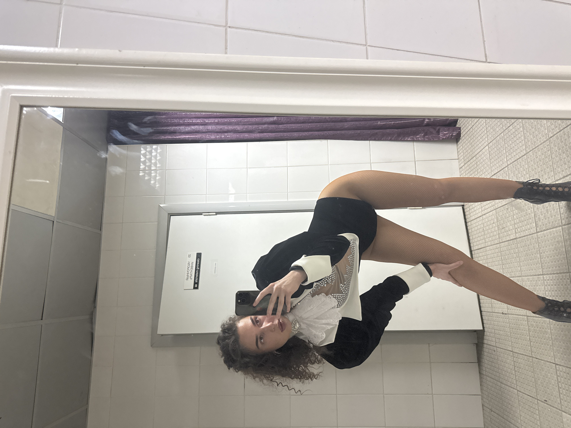 A person is taking a selfie in a mirror. The person has curly brown hair and is wearing a black jacket with white cuffs, a white ruffled collar with a jeweled brooch, and fishnet stockings. They are also wearing black heeled boots. The background appears to be a tiled room, possibly a dressing room or backstage area, with a door that has a sign in Russian. There is a purple sequined curtain visible in the mirror reflection. The ceiling is made of acoustic tiles. The floor is tiled with a grid pattern. The overall impression is that of a performance or event setting.