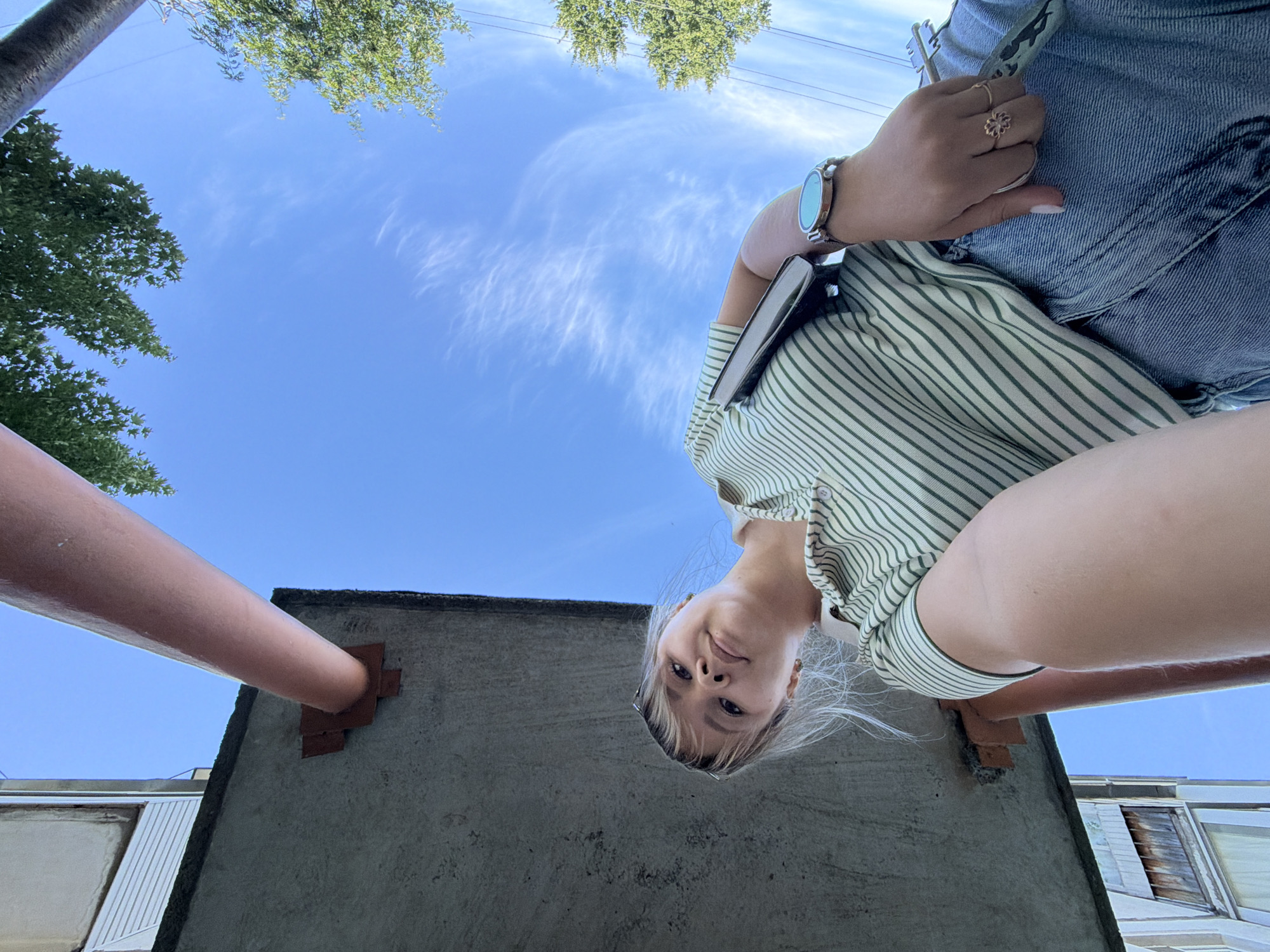 A young woman with blonde hair tied in a bun is photographed from a low angle looking up at the sky, with the underside of a concrete structure and a reddish pipe in the foreground. She is wearing a green and white striped polo shirt and denim jeans. Her left arm is extended towards the camera, holding a dark-colored phone, and her right hand, adorned with rings, rests on her hip. A wristwatch with a light blue face is visible on her right wrist. The background consists of a clear blue sky with wispy clouds and green foliage from trees. The concrete structure appears to be part of an outdoor urban setting, possibly a balcony or walkway, with visible rust stains and mounting brackets. The lighting suggests it is daytime. The overall scene has a casual, outdoor ambiance.