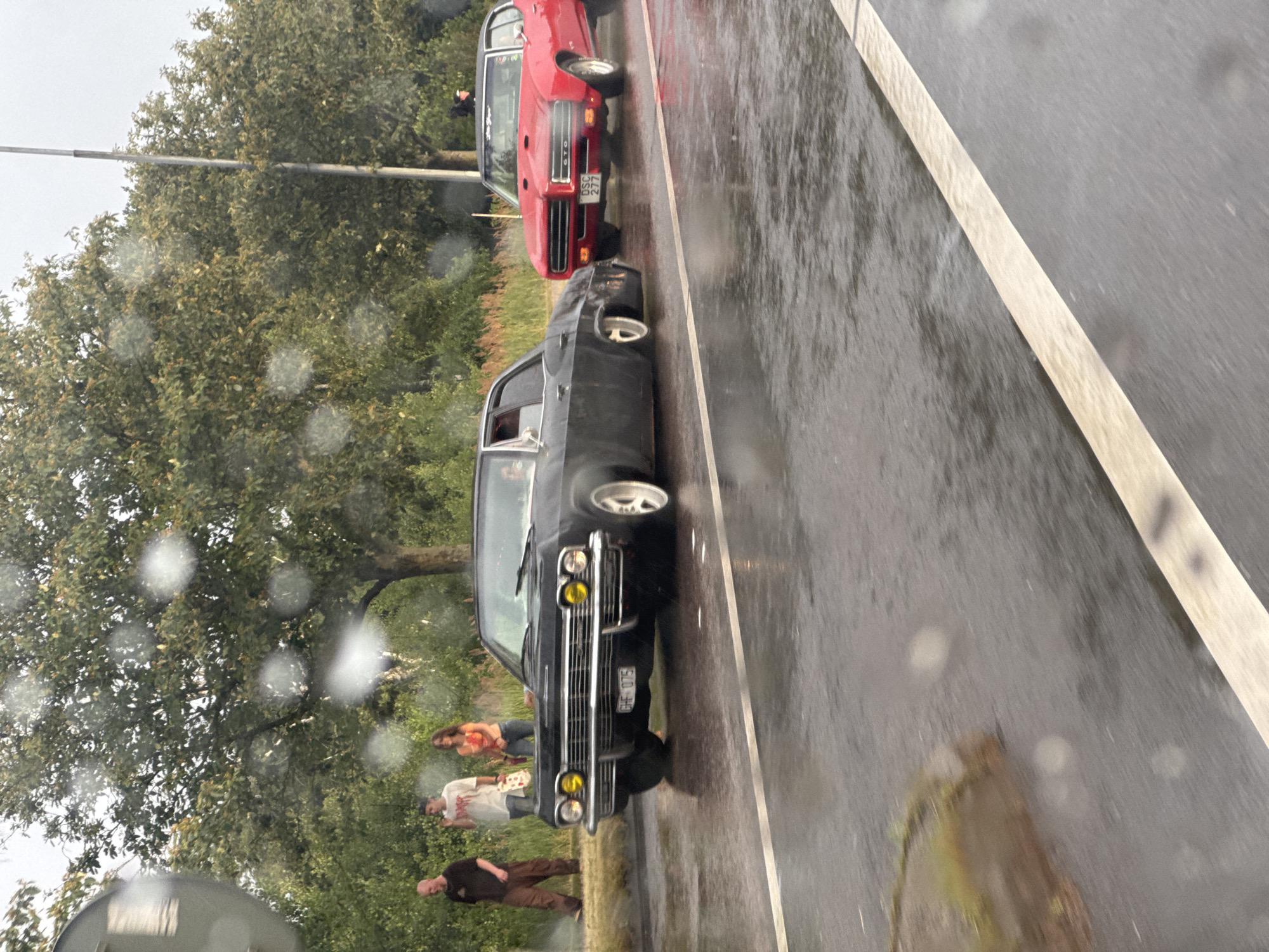 In Falkenberg, Sweden, on a wet road, two vintage American cars are parked. The car in the foreground is a black, four-door sedan with yellow headlights. It has a license plate that reads "CHF 075". Behind it, to the right, is a red two-door coupe with the license plate "DSC 277". The red car appears to be a Pontiac GTO. Three people are standing on the sidewalk to the left of the black car. One is a man in a dark shirt and brown pants, another is a man in a white t-shirt and jeans, and the third is a woman in an orange and white top and jeans. The trees in the background are full of green leaves, and the sky is overcast, suggesting it might be raining or has recently rained. The road surface is wet and reflective, indicating damp conditions.