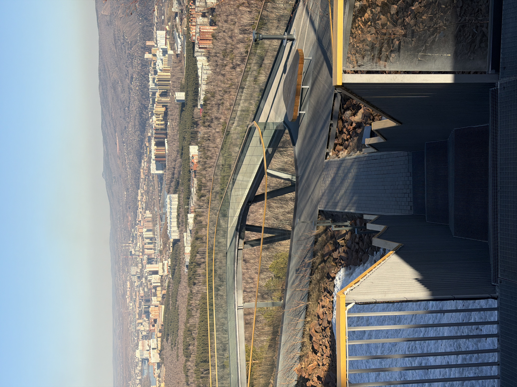 The image shows a panoramic view of Krasnoyarsk, Russia, from the observation deck of the Torgashinsky Ridge. A pedestrian bridge with glass railings extends from the deck, overlooking the city and surrounding forested areas. The city skyline includes residential and commercial buildings, nestled between dense rows of trees. The sky is clear and pale blue, with a slight haze. In the foreground, the observation deck features architectural elements like metallic structures, walkways, and rock arrangements. Some areas show remnants of snow or ice. The image is taken from an elevated position, emphasizing the height and perspective of the vantage point.