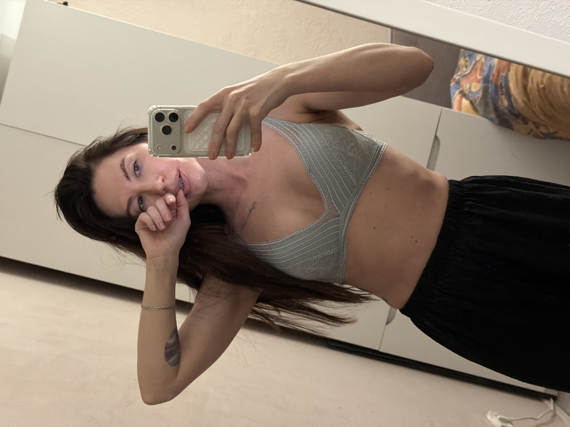 A woman is taking a selfie in a room with white furniture. She is wearing a light blue bra and black pants. She has dark hair and is holding her phone in her right hand. Her left hand is near her mouth. There is a tattoo on her left forearm. The location is not discernible from the image, so it is impossible to verify it is in Perm, Russia.