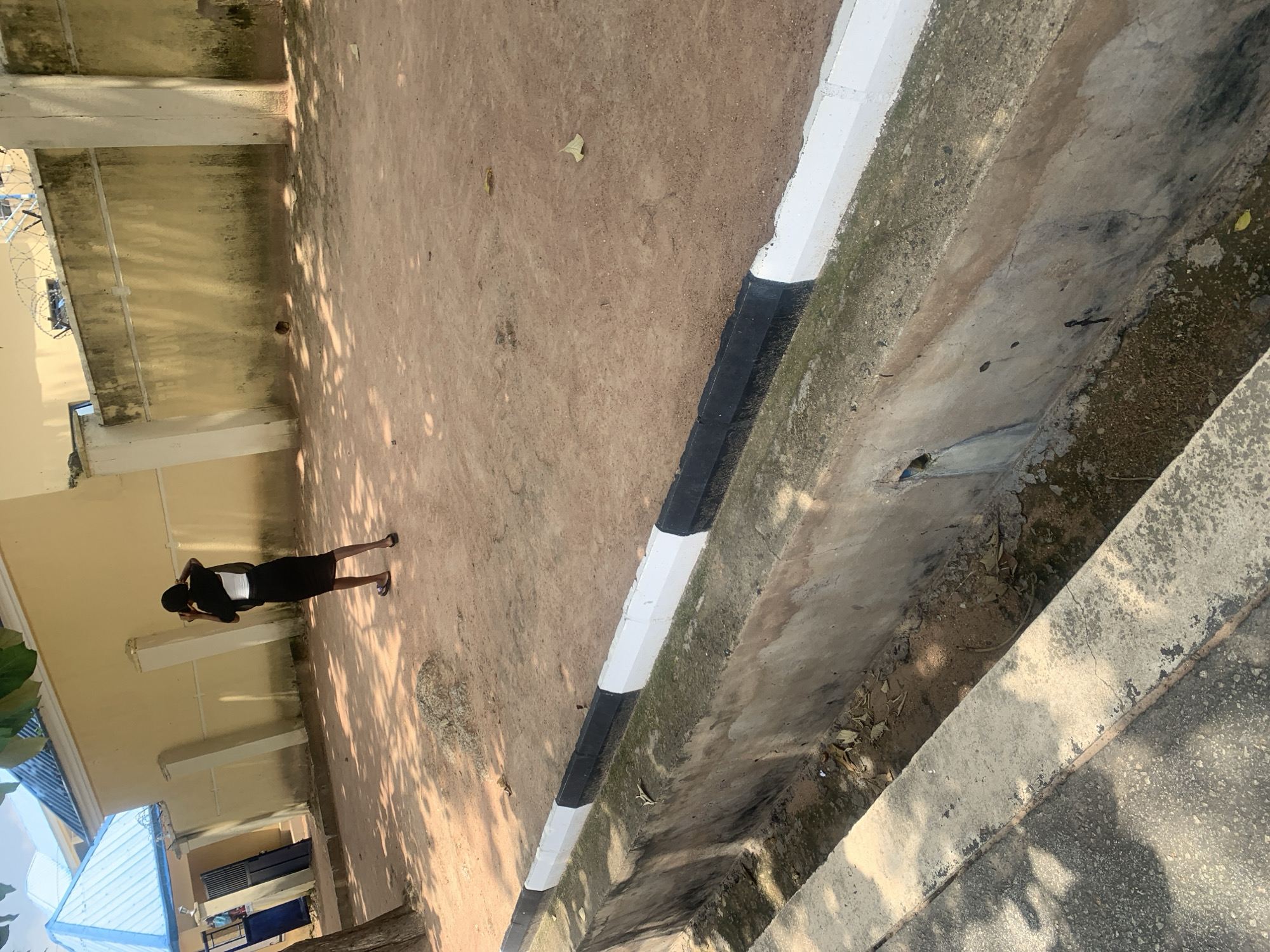 In Jalingo, Nigeria, a young woman stands in a dusty courtyard, facing away from the camera and holding a phone to her ear. She wears a black dress and appears to be in motion, with her left leg slightly forward. The courtyard is bordered by a drainage channel with a black and white painted curb. Behind her, a light yellow building with a blue metal roof stands, and a section of a concrete wall topped with barbed wire can be seen to the right. The ground is covered in reddish-brown soil, scattered with a few leaves and small stones. Sunlight casts shadows across the scene, indicating it is daytime.