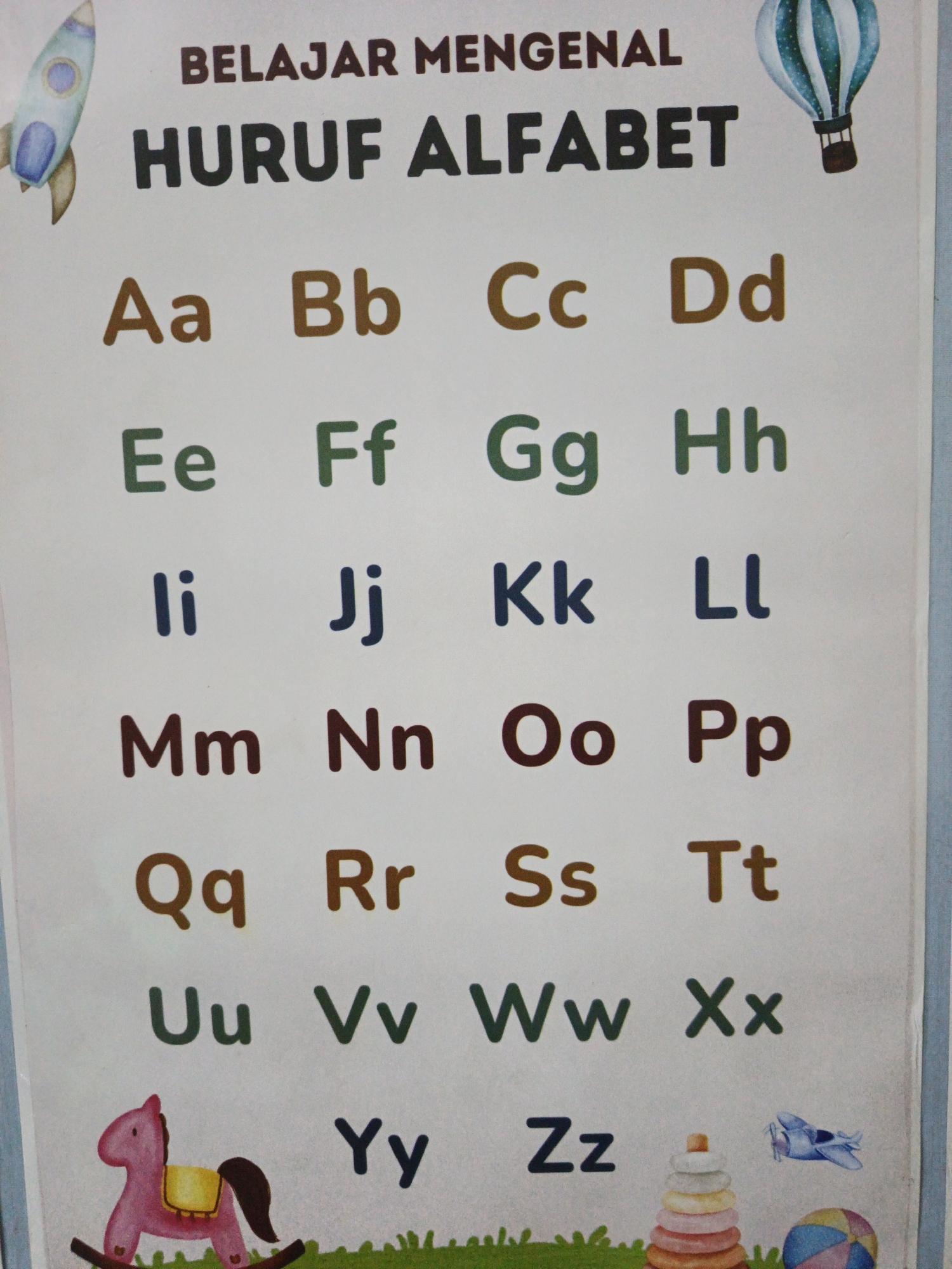 The image displays an alphabet chart with Indonesian text at the top that reads "BELAJAR MENGENAL HURUF ABALABET". The chart presents capital and lowercase letters of the alphabet arranged in rows. Each letter is colored differently, with combinations of brown, green, and blue. Decorative illustrations are present at the top and bottom of the chart, including a rocket, a hot air balloon, a rocking horse, a toy airplane, a stacking toy, and a ball. The chart is hung on a light-colored wall. The scene does not provide any information about the city of South Jakarta, Indonesia, or any specific location within it. There are no people or actions depicted in the image.