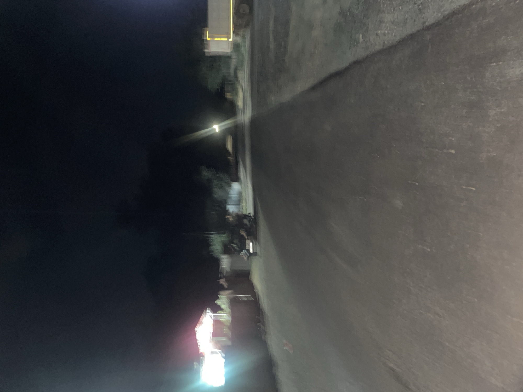 This is a night-time, horizontal, medium-shot photograph of a paved road in Ak-Jol, Kyrgyzstan. The road appears to be under construction or repair, as evidenced by the presence of construction vehicles and equipment.
In the background, on the left side of the image, there is a brightly lit building with a red-tiled roof. Next to the building, a dark-colored construction vehicle, possibly a grader or roller, is positioned on the road. Further down the road, on the right side, a large yellow dump truck with its bed raised is parked.
The road itself is dark asphalt, with some areas appearing freshly laid. The environment is dark and sparse, with trees and vegetation visible on the sides of the road. A single streetlamp illuminates a portion of the road in the distance. There are no visible people or animals in the frame, and the overall impression is one of a quiet, industrial setting at night.
