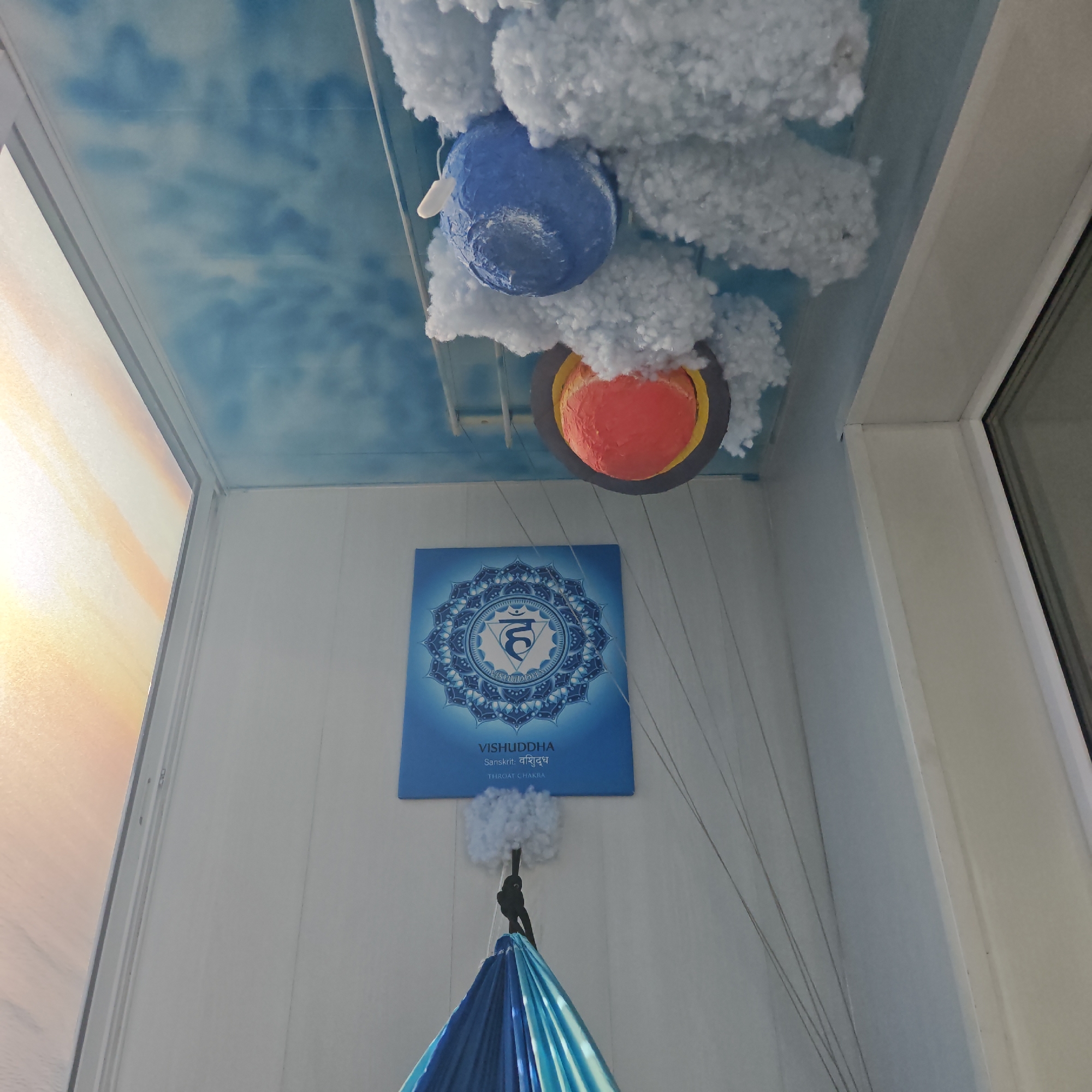 The image depicts an interior space, possibly a child's room, in Rostov, Russia. The ceiling is painted with a cloudy blue sky motif, from which hang artificial clouds and rudimentary models of planets. Below, on a white wall, is a poster depicting the Vishuddha chakra symbol with the words "VISHUDDHA, Sanskrit: विशुद्ध, THROAT CHAKRA." Suspended beneath the poster, and attached to it with white fluffy material, is a blue fabric hammock or swing. Fine wires or strings run from the ceiling towards the floor. The room is illuminated by light entering through windows on either side of the frame. The overall impression is a space designed for relaxation and contemplation, potentially related to yoga or spiritual practices.