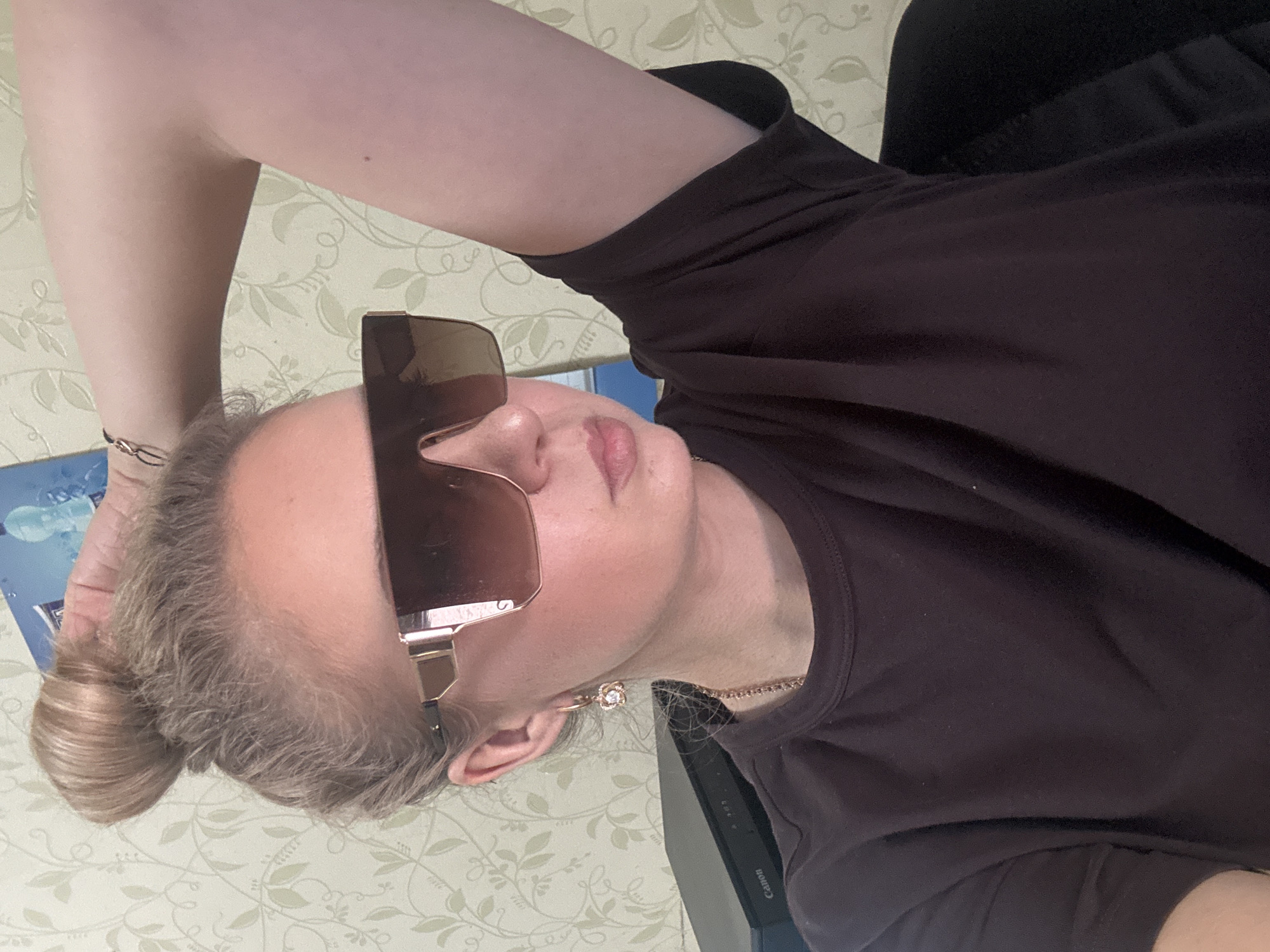 A young woman with blonde hair pulled into a messy bun poses for a selfie, wearing large, dark, angular sunglasses. She is dressed in a dark brown t-shirt and has on a delicate gold necklace with a pendant, and a flower-shaped earring in her left ear. Her left arm is raised behind her head, and her right arm is out of frame. The background is a beige wall with a subtle floral pattern. To the left of the woman, part of a black electronic device with the word "Canon" visible on it is in the frame. The lighting is soft and appears to be coming from the front.