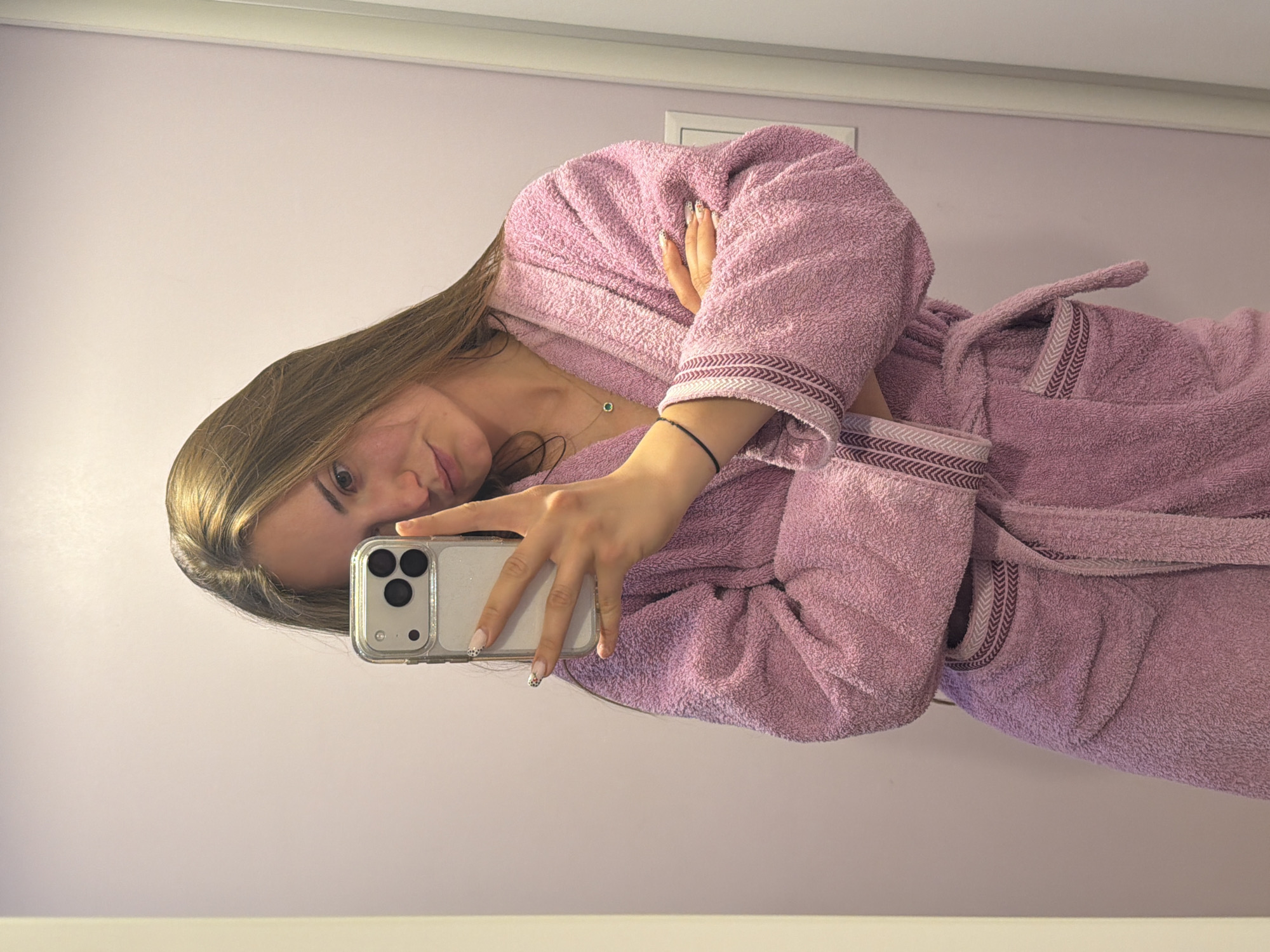 A woman wearing a pink bathrobe is taking a selfie with her smartphone. She is indoors, against a light purple wall with a white border. The location is not explicitly depicted to be in Buhovo, Bulgaria.