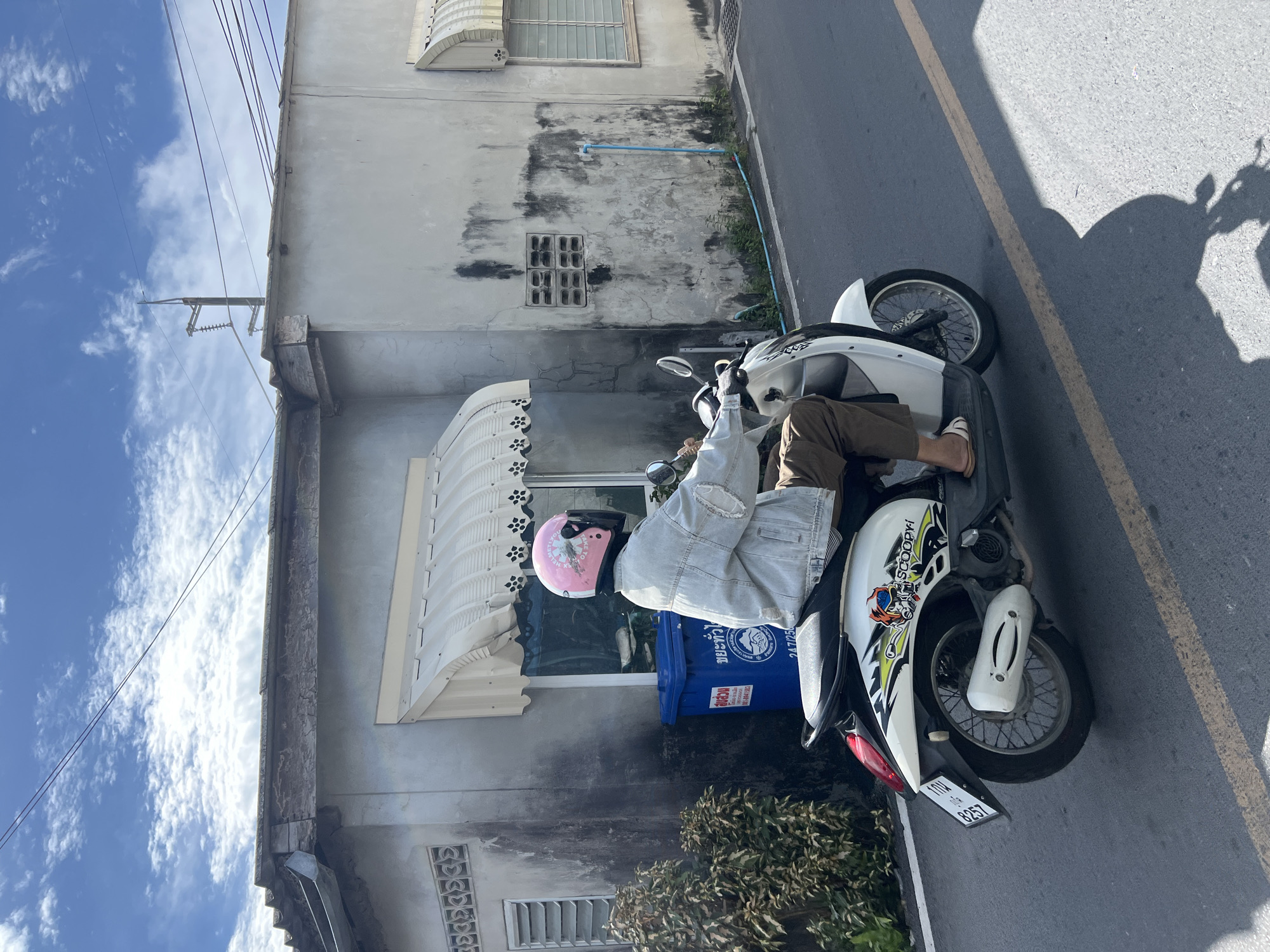A person wearing a light blue denim jacket, brown pants, and white sandals is riding a white scooter with graphics on the side, including the word "SCOOPY-I". They are wearing a pink helmet with floral stickers and a dark visor. The scooter has a Thai license plate that reads "10W 8257". The person is on a paved road with a yellow line marking. To the left of the road is a sidewalk with a green bush and a blue trash bin with Thai writing. Behind the trash bin is a building with a gray, weathered facade, a white awning, and a window with blinds. To the right of the building is a street with a blue pipe running along the ground and another building with a similar weathered facade and an awning. The sky above is blue with scattered white clouds. The scene appears to be in a residential or commercial area in Choeng Thale, Thailand.