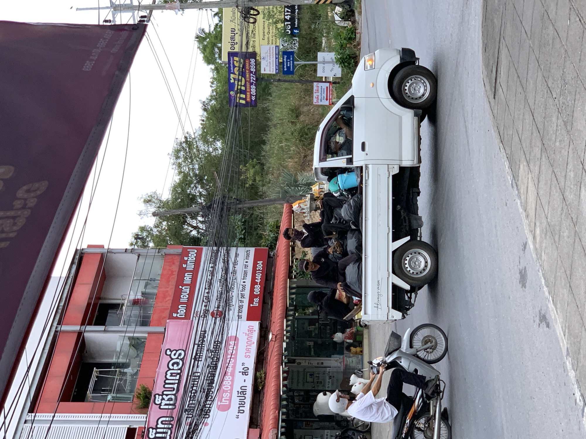 In the city of Ko Samui District, Thailand, a white pickup truck is parked on the right side of a street. The truck is carrying several people and large black bags in its bed. The people appear to be wearing dark clothing and some have their faces obscured. In the foreground, a person on a silver motorcycle is riding past the truck on the left. The motorcycle rider is wearing a white helmet and dark clothing. The street is paved and there is a sidewalk on the right. In the background, there are buildings with red awnings and signs in Thai. Trees and utility poles are also visible. The scene appears to be a busy street in a commercial area.