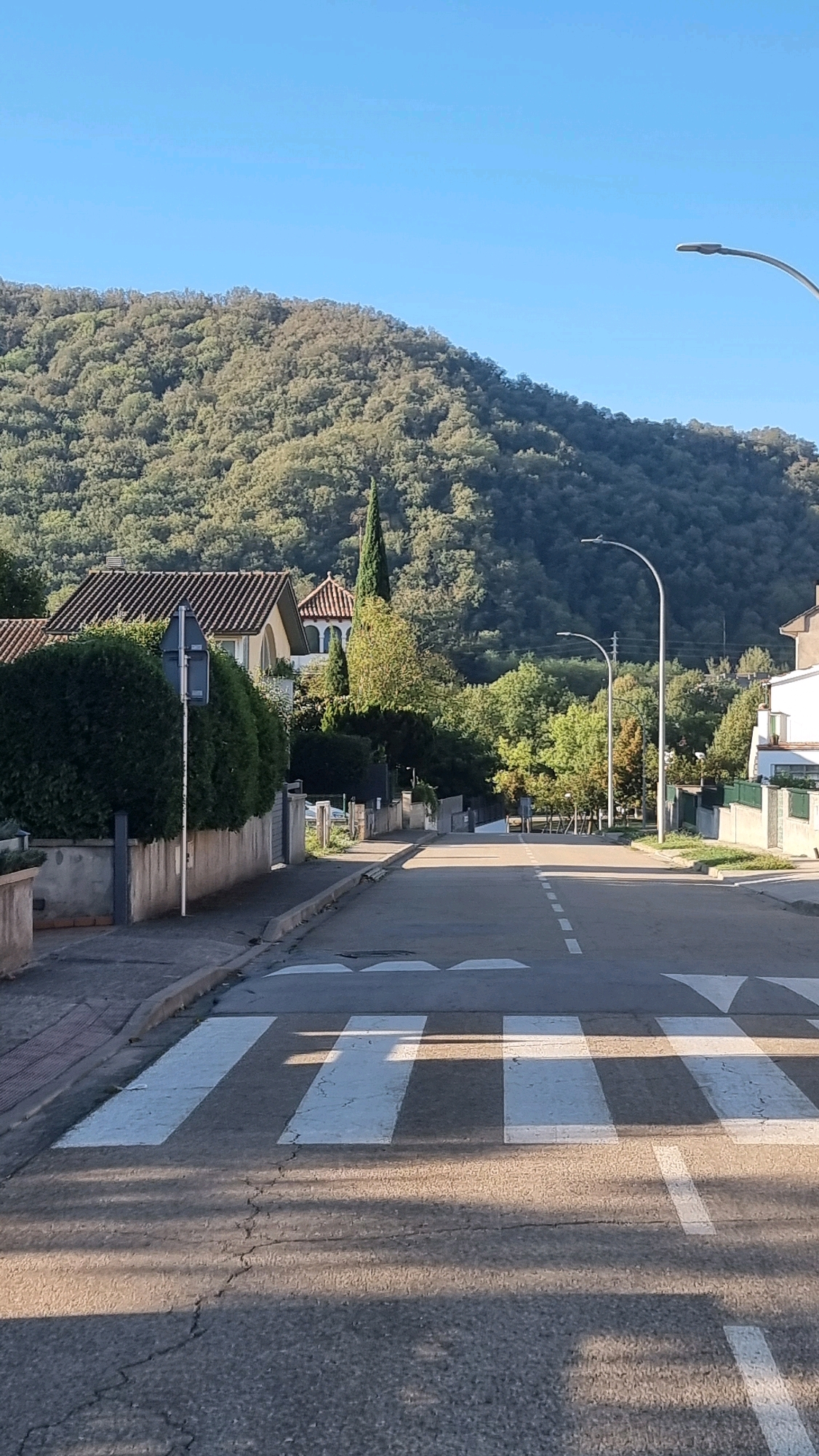 The image depicts a street scene in Sant Joan Les Fonts, Spain, with a pedestrian crossing in the foreground. A paved road stretches uphill, lined with residential buildings and lush greenery. A steep, tree-covered hill dominates the background. The sky is clear and blue. A streetlamp is visible on the right side of the road. The road surface shows cracks and is marked with white lines indicating lanes and the pedestrian crossing.