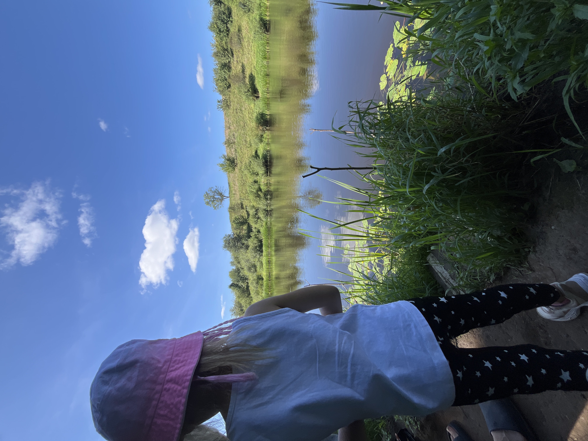 In a serene natural setting, a young child wearing a pink bucket hat and a white tank top stands facing a body of water. The child's black leggings are adorned with white star patterns. The surrounding environment features lush green grass and vegetation along the water's edge, with lily pads visible on the surface of the calm river. Across the water, a gently sloping grassy hill is dotted with trees and shrubs under a bright blue sky with scattered white clouds. The scene captures a peaceful moment outdoors, possibly near the village of Germanovichi within the Germanovichi rural council of Belarus.