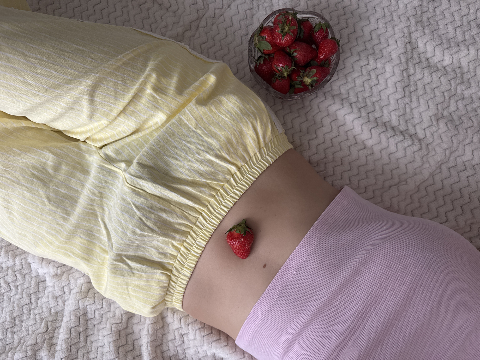 A high-angle, close-up shot shows a person lying on a textured white blanket. The person wears yellow and white striped pants and a light pink shirt. A single strawberry sits on the person's stomach. A glass bowl filled with strawberries is positioned near the person's waist. The photograph does not contain sufficient information to determine if the subject lives in Izhevsk, Russia.