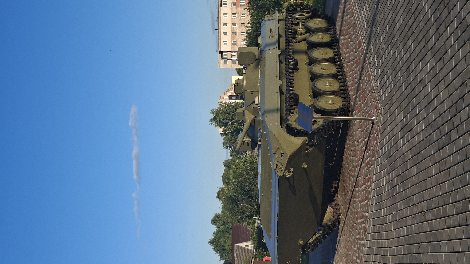 A green military tracked vehicle is displayed on a paved area in front of trees and residential buildings under a clear blue sky. The vehicle, likely an amphibious infantry fighting vehicle or armored personnel carrier, has a long, flat front designed for water traversal. It features a turret with a gun, a series of road wheels along its tracks, and is positioned on a brick-patterned pavement with some reddish-brown accents. A simple metal stand with a blue plaque is located in front of the vehicle. The background shows a line of trees and multi-story apartment buildings, suggesting an urban or suburban setting. The lighting indicates it is daytime, likely morning or late afternoon, given the long shadows. The city of Smalyavichy, Belarus is implied as the location.