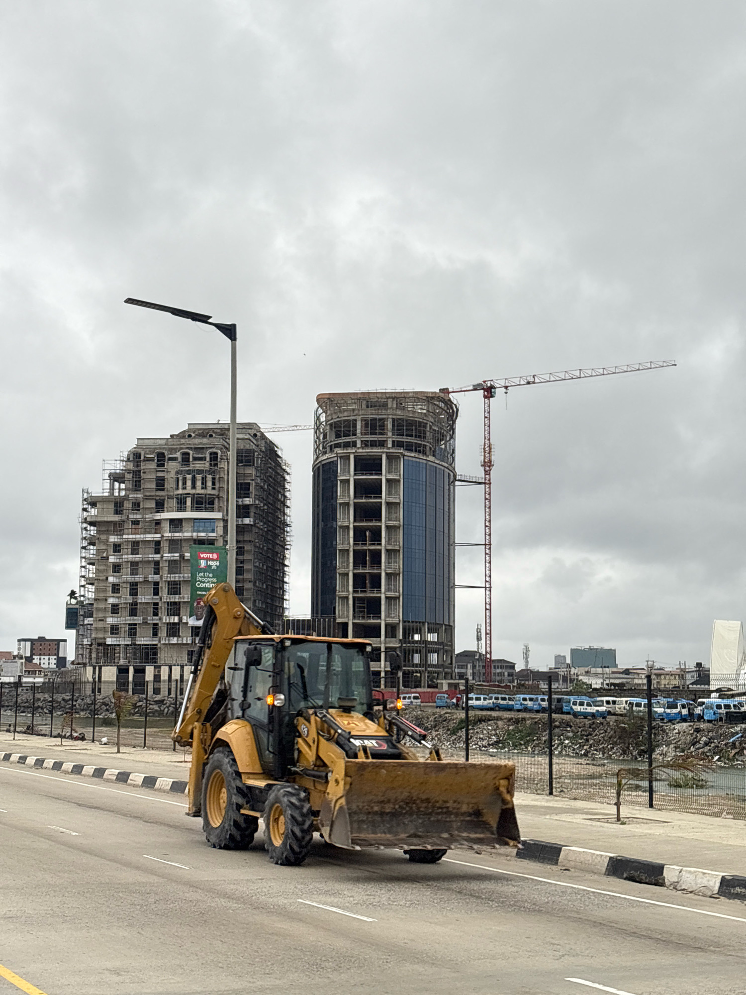 This is a wide-angle, eye-level shot of a construction scene in Ilado, Nigeria, under a heavily overcast sky. In the foreground, a yellow Caterpillar backhoe loader is driving on a paved road with white and black painted curbs. The machine's front loader is slightly raised, and its tires are large and knobby.
Behind the backhoe, a large building, covered in scaffolding, is under construction. It has a dark-colored facade and appears to be several stories high, with visible window openings. To the right of this building, another taller, more modern-looking skyscraper is in the process of being built, featuring a glass facade and a skeletal concrete structure at its upper levels. A red construction crane extends from behind this building and into the sky.
Further in the background, a row of blue and white minibuses are parked along a fenced area. Beyond the minibuses, other buildings of varying heights and architectural styles can be seen, suggesting a bustling urban environment. The overall impression is one of ongoing development and construction within the city.